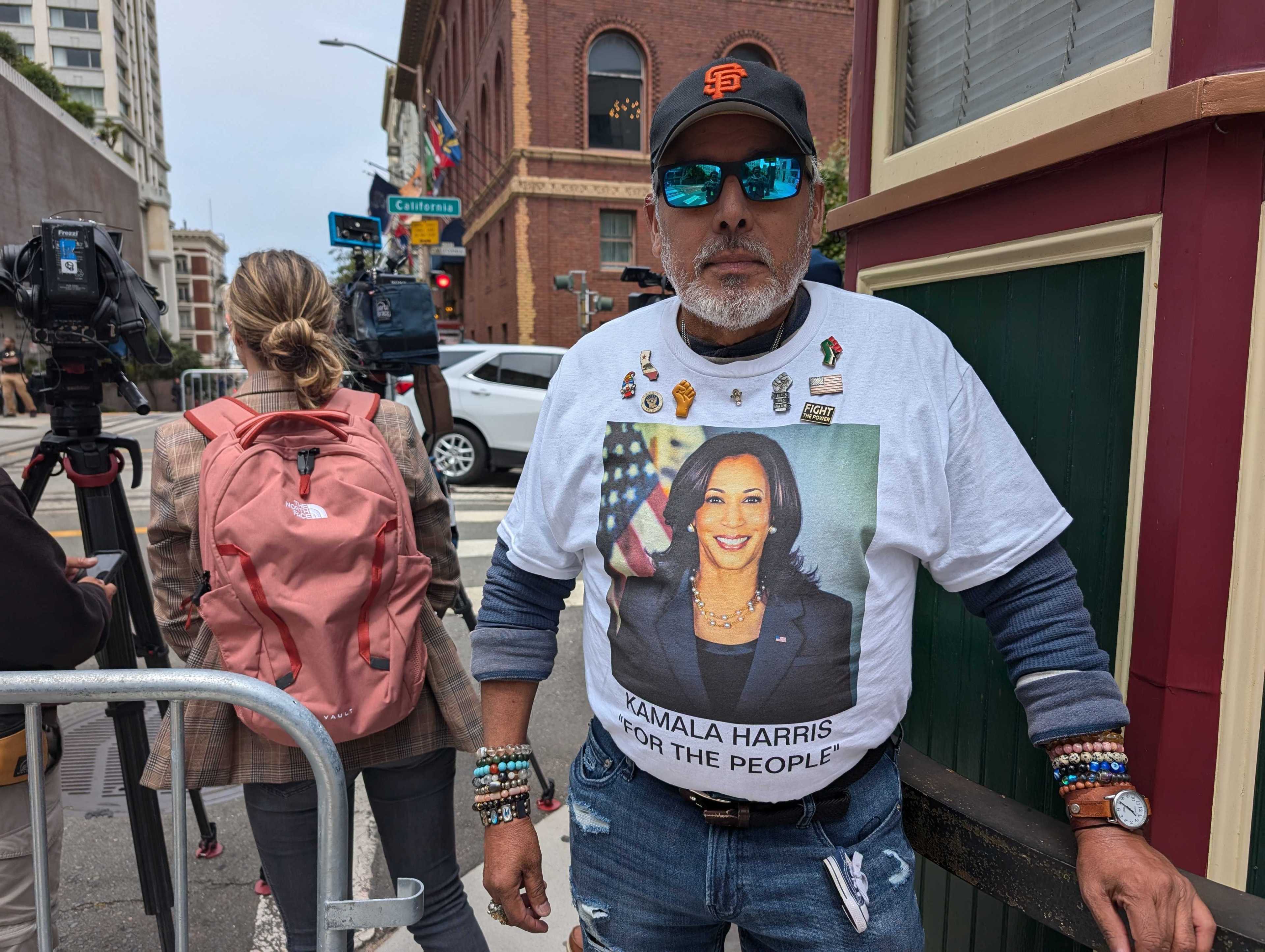 A bearded man in sunglasses wears a shirt with Kamala Harris' photo among a small crowd, including a woman in a pink backpack and videographers on a street corner.