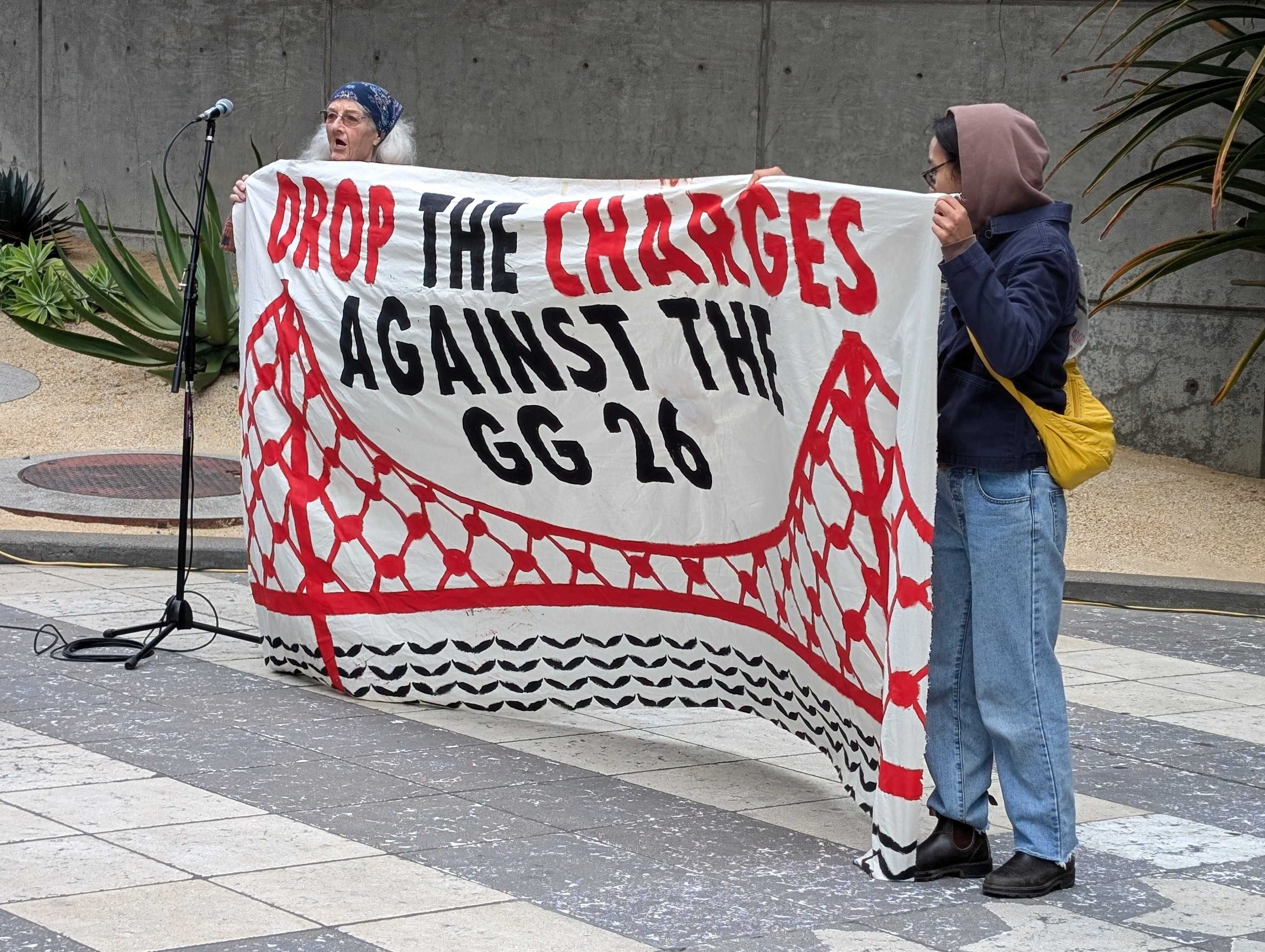 Two people hold a banner that says "drop the charges against the GG 26," referring to the 26 people facing charges from a demonstration that blocked traffic on the Golden Gate Bridge in April.