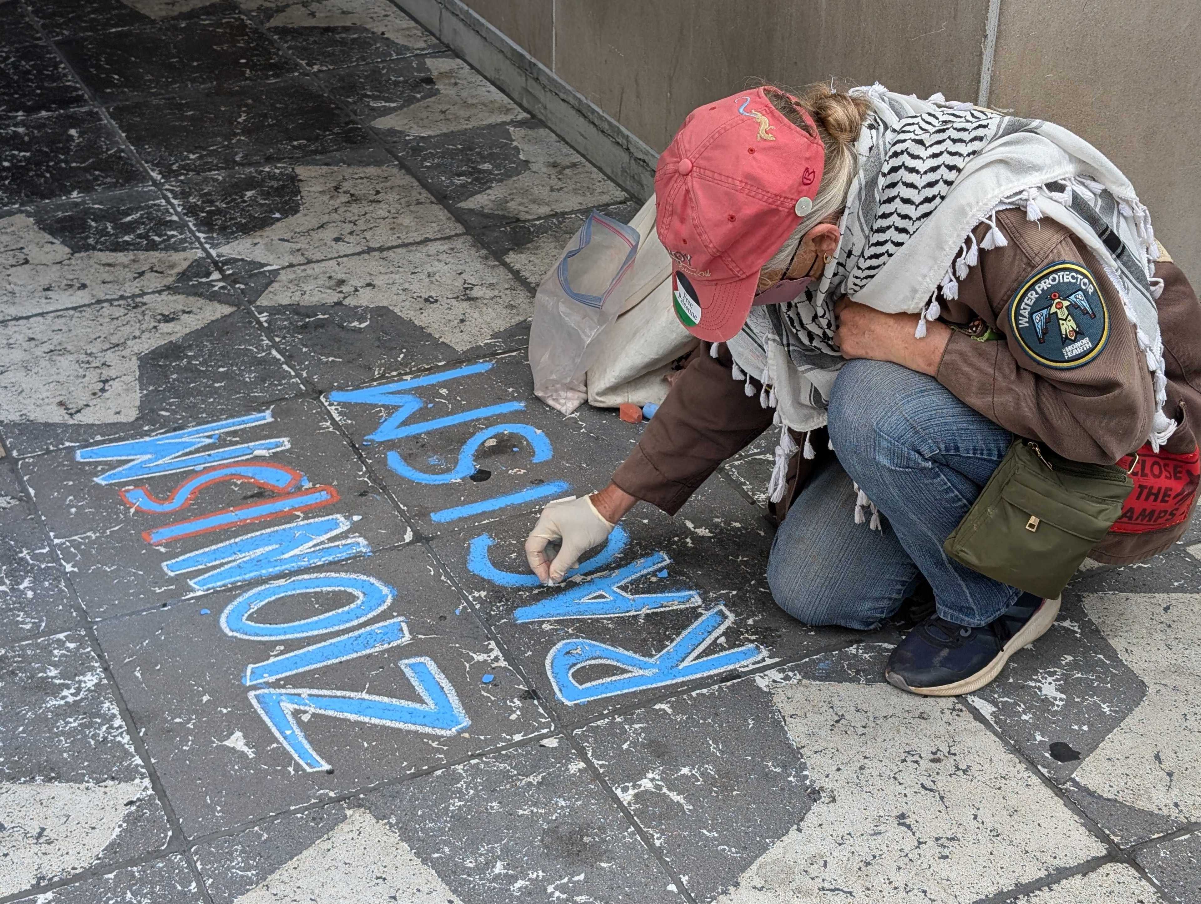 A person in a red baseball cap writes that &quot;Zionism is racism&quot; in chalk on a sidewalk.