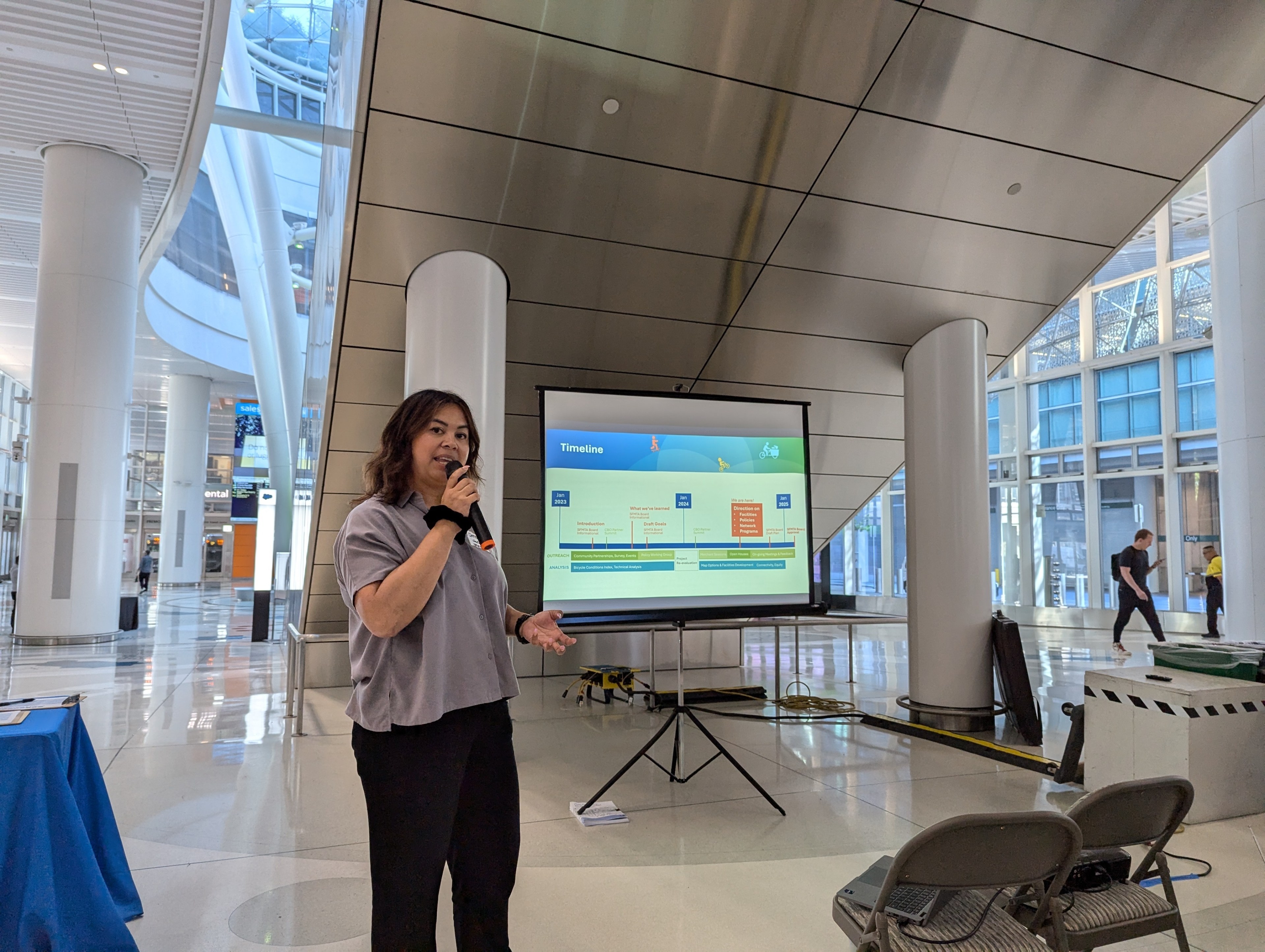 A woman is speaking into a microphone beside a screen displaying a timeline. They are in a modern, spacious area with large pillars and glass windows.