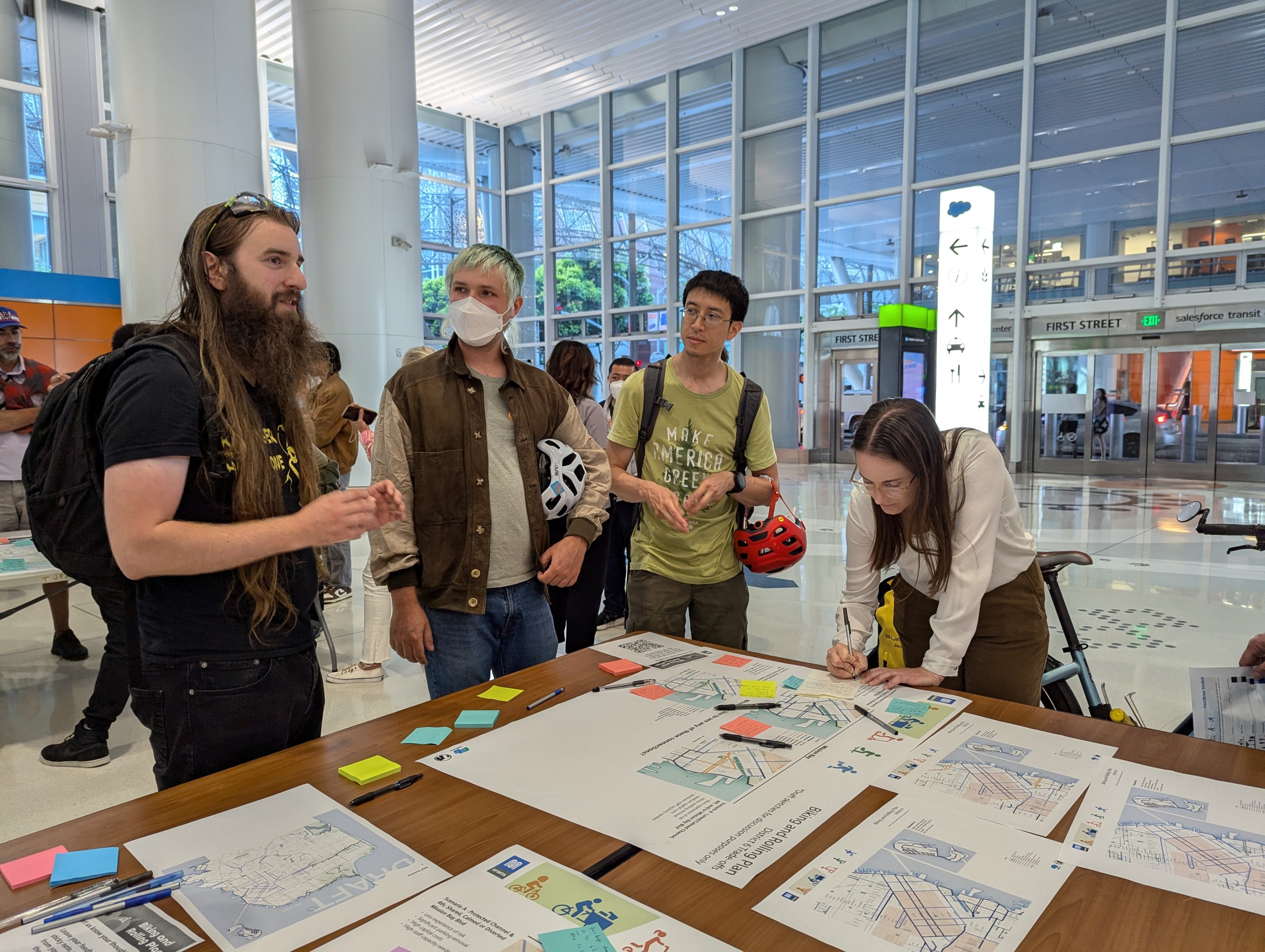 A group of people are in a modern indoor space discussing maps spread out on a table; one is writing notes, while others listen and engage in conversation.