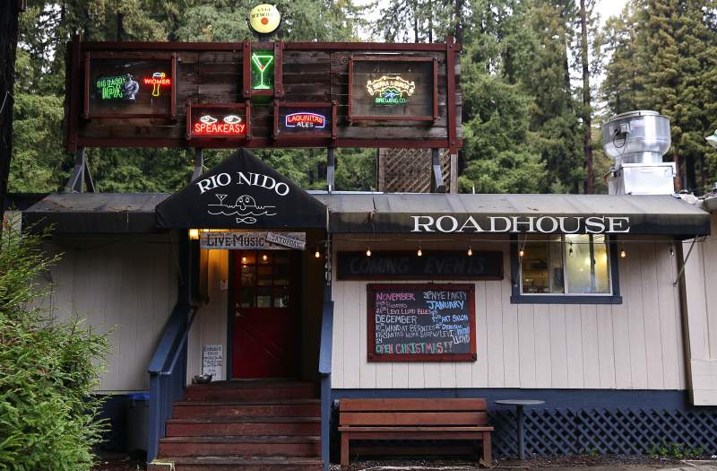 The image shows the exterior of Rio Nido Roadhouse, featuring neon signs, a black awning, stairs leading to a red door, a bench, and a chalkboard listing events.