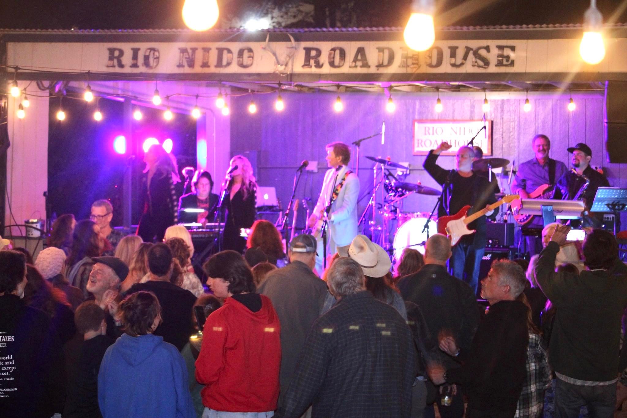 A lively band plays on an outdoor stage at night under string lights at the Rio Nido Roadhouse, entertaining a crowd of people dancing and enjoying the music.