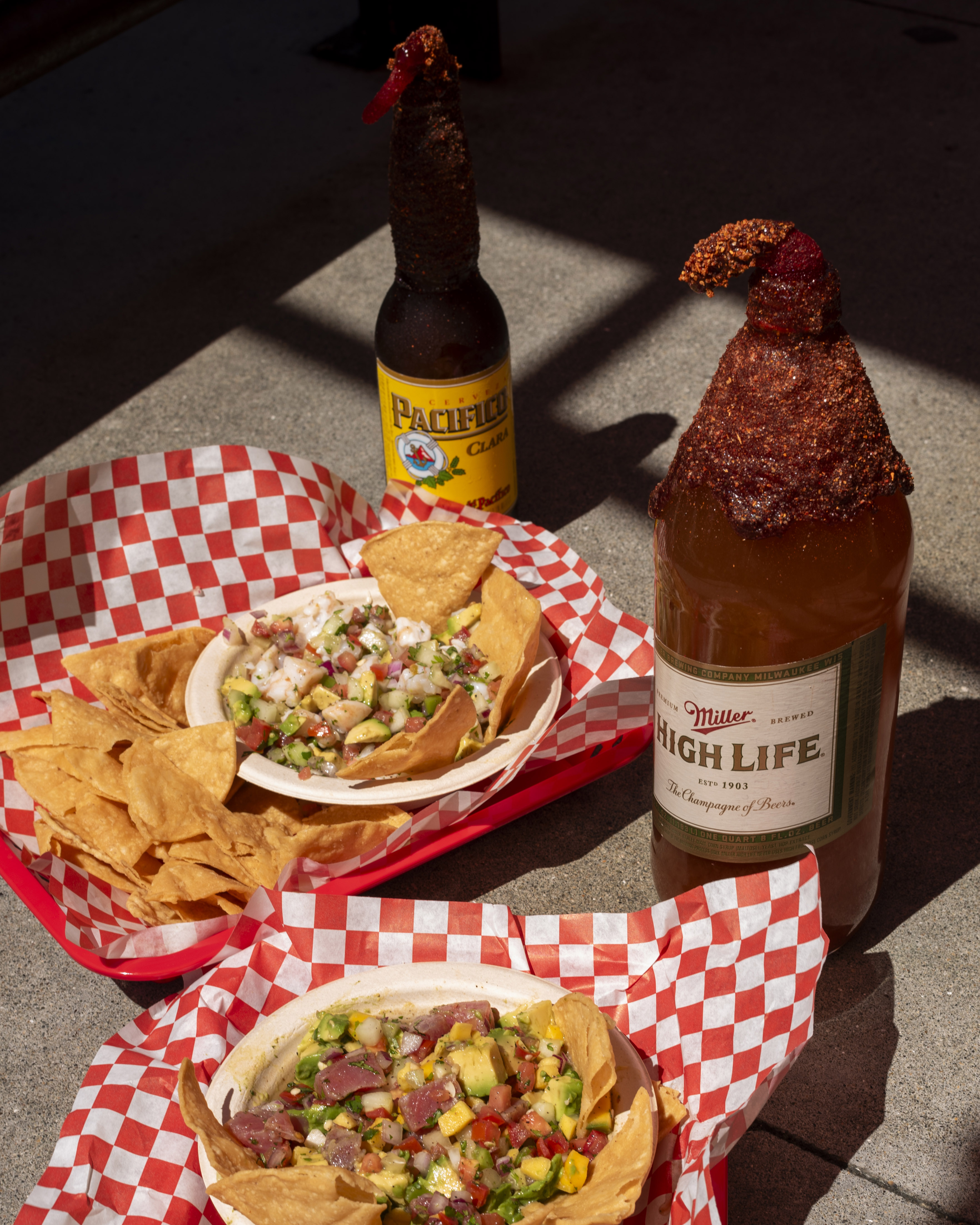 Two beers with spicy rimmed tops are next to baskets of tortilla chips and dips, displayed on checkered red and white paper trays.