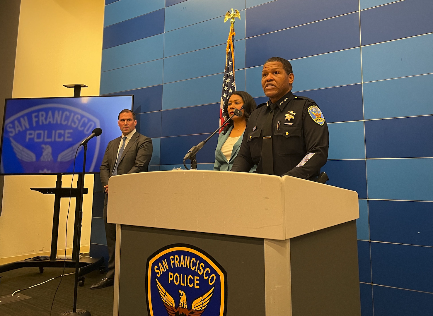 A policeman speaks at a San Francisco Police podium, accompanied by two individuals. An American flag and a large monitor displaying the police emblem are behind them.