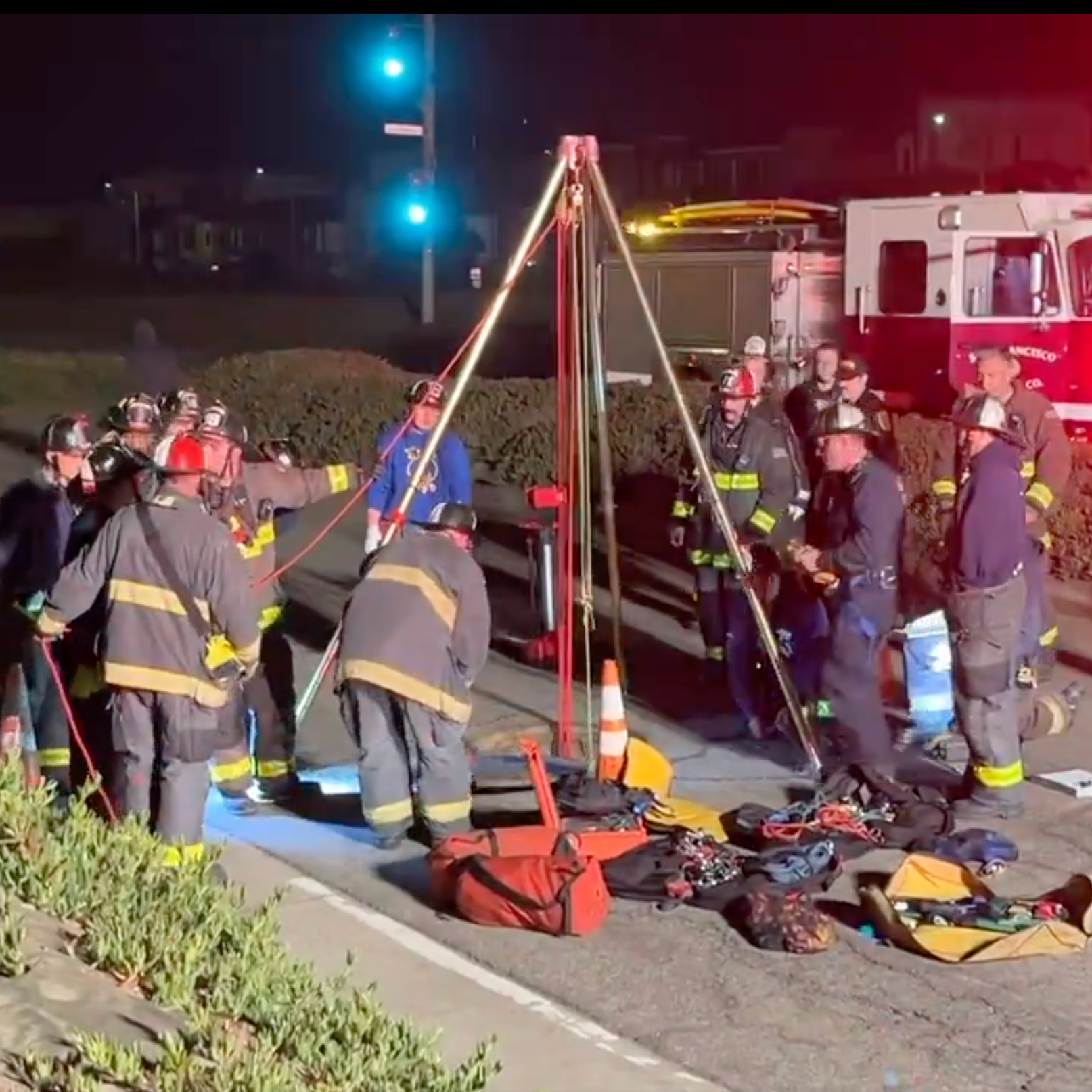 Firefighters are gathered around a tripod setup with ropes and equipment on the roadside at night, illuminated by emergency vehicle lights in the background.