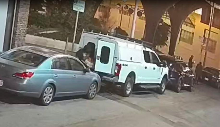 A person appears to be breaking into a parked white truck at night in a residential area, with other cars and a couple of people visible in the background.