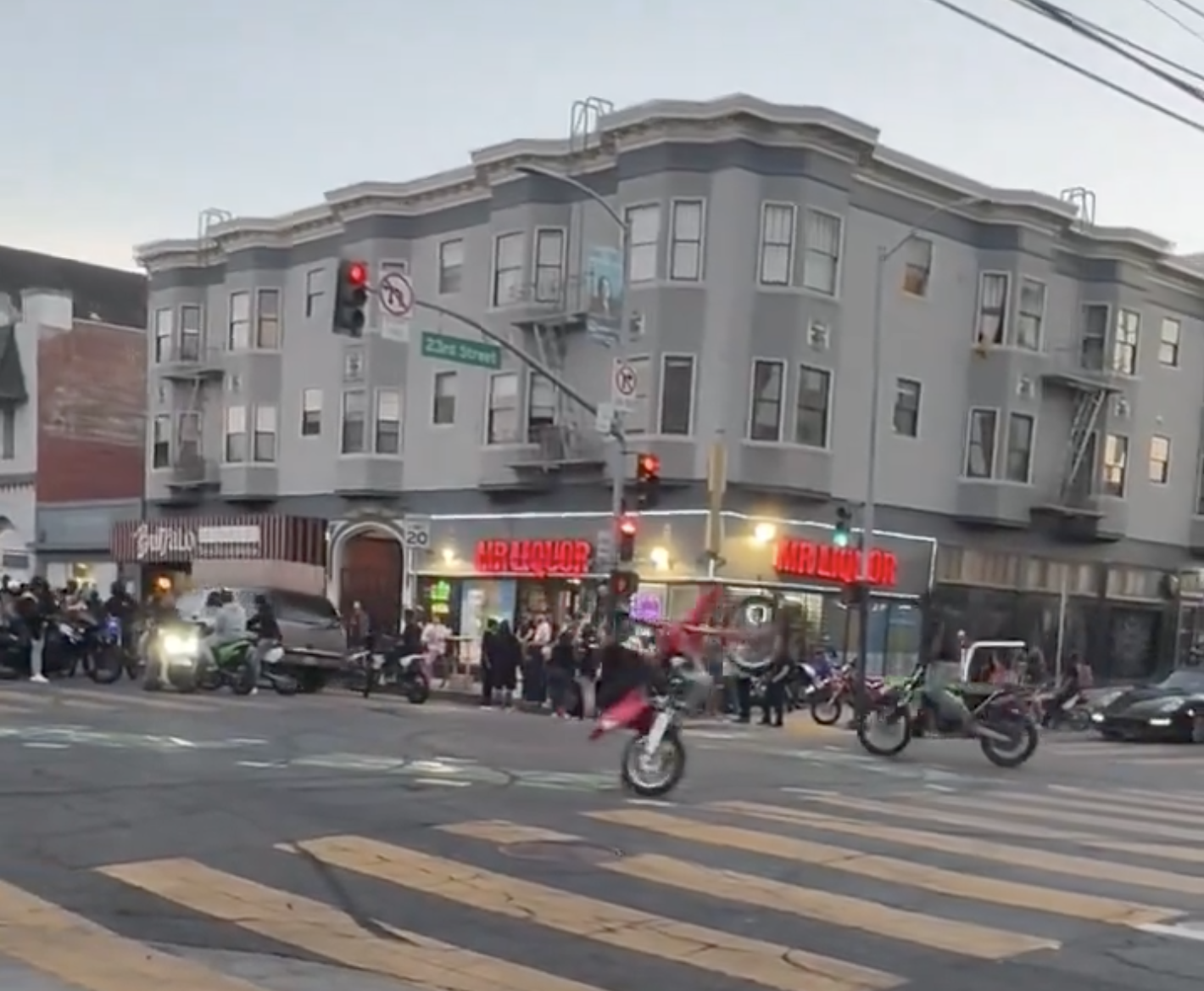 A bustling street corner with people gathered, motorcycle riders performing stunts, a grey building with a bar and liquor store, and traffic lights glowing red.