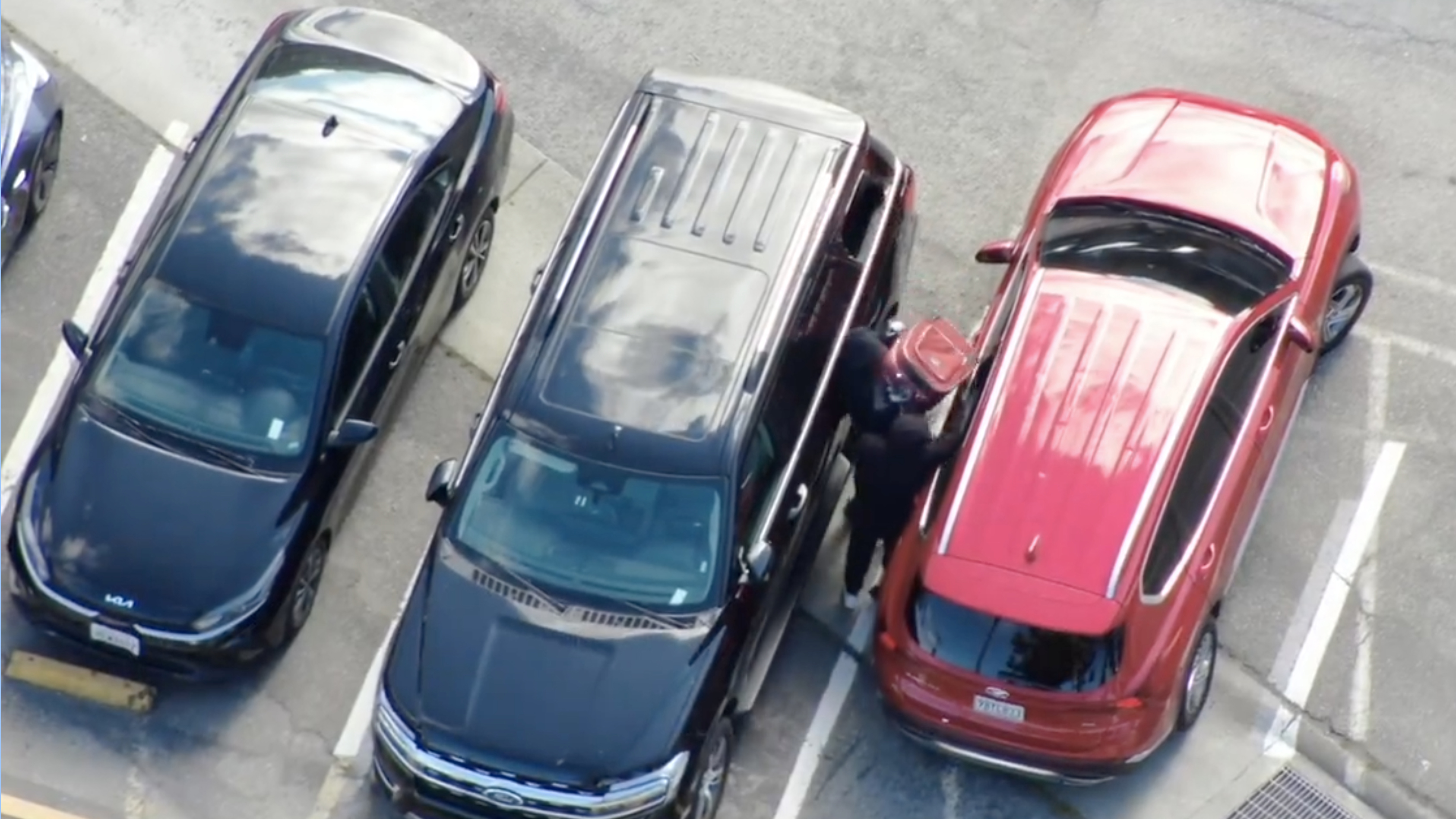 The image shows three cars parked closely together in a parking lot: two black cars on the left and a red SUV on the right with a person between the black SUV and the red car.