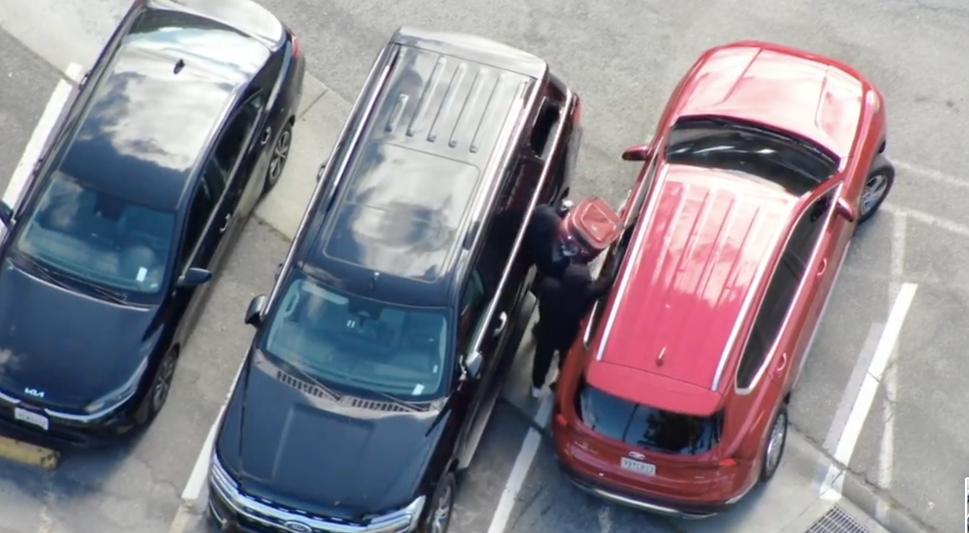 In a parking lot, a person stands between a red SUV and a black vehicle, appearing to break into the red car. A third black car is parked beside them.