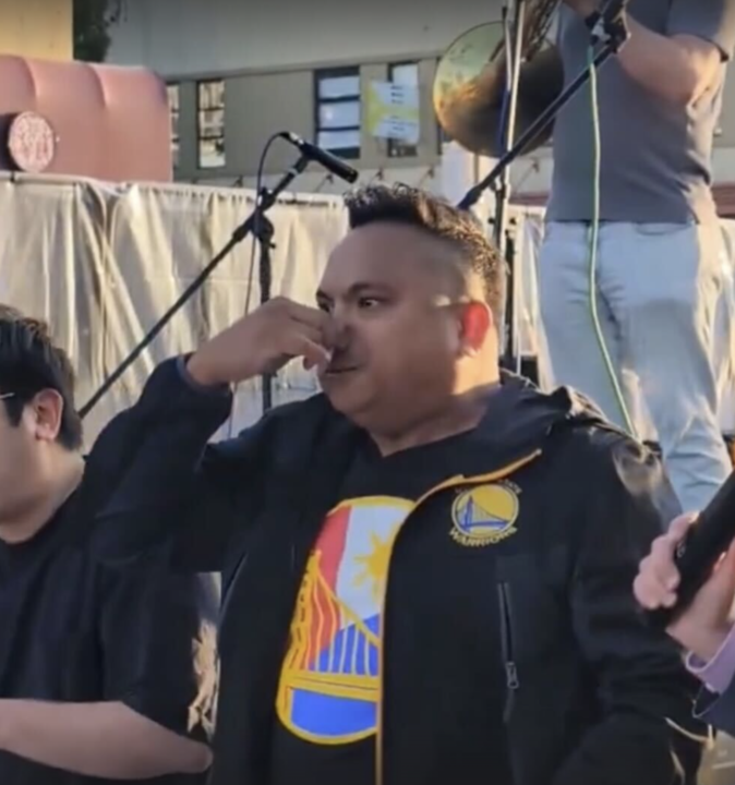 A man in a black and yellow jacket holds his nose while standing on a stage, with a microphone stand and another person holding a microphone behind him.