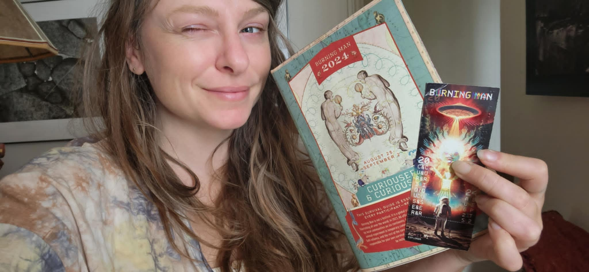 A woman smiles and winks at the camera, holding a Burning Man 2024 guide and a colorful event ticket in her hands.