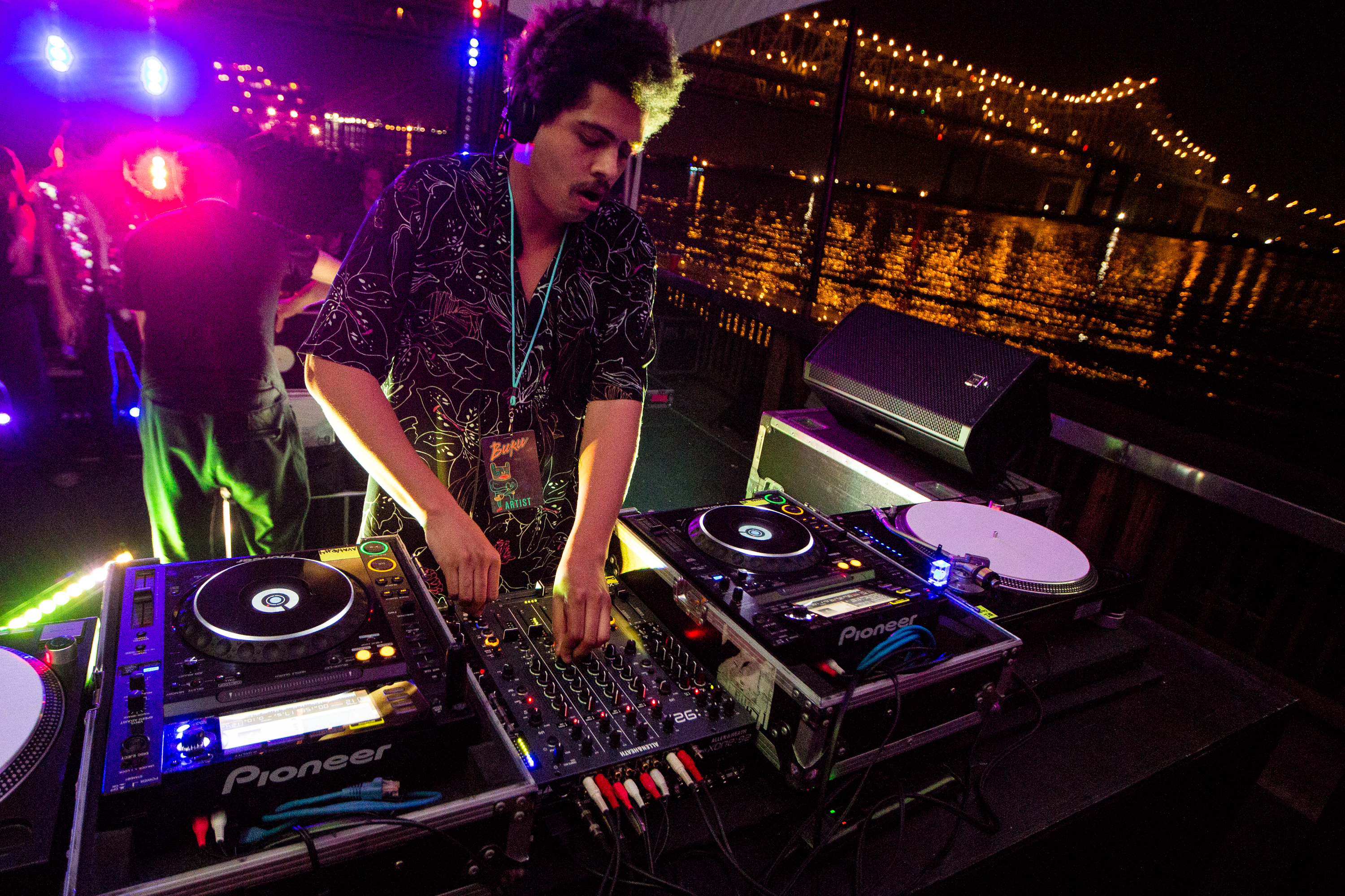 A DJ, wearing headphones and a name tag, mixes music on a deck with colorful lights, a cityscape, and a bridge with bright reflections over the water at night.