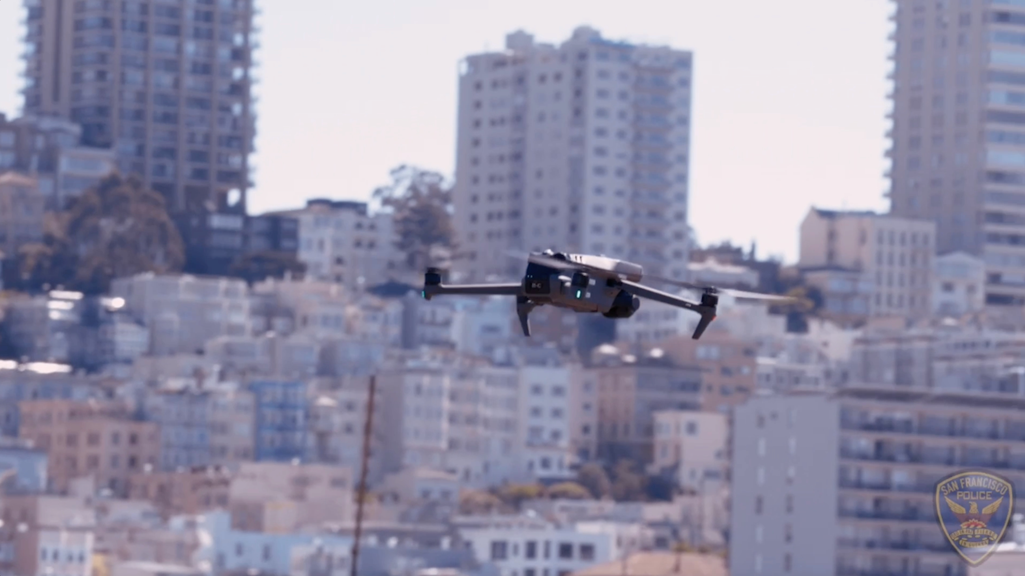 A drone is flying in front of a densely packed urban area with numerous tall buildings, possibly in San Francisco, as suggested by a visible police badge.