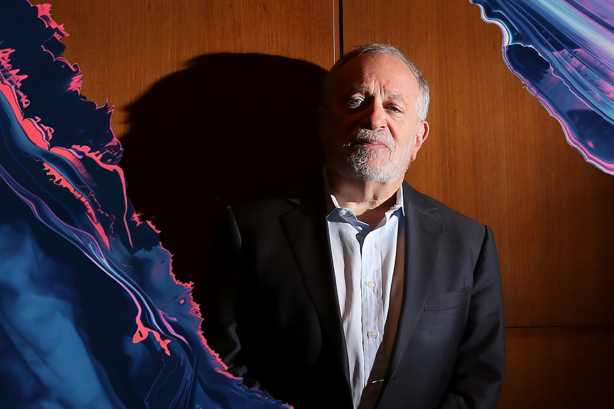 Robert Reich, in a dark suit and white shirt, stands against a wooden wall with abstract blue and pink shapes on the left and right edges.