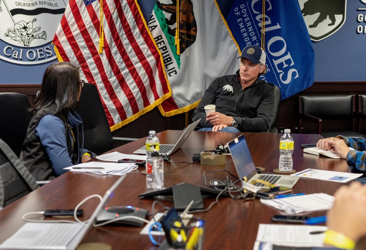 Several people sit around a conference table with laptops, documents, and water bottles. U.S. and California flags are displayed on the wall behind them.