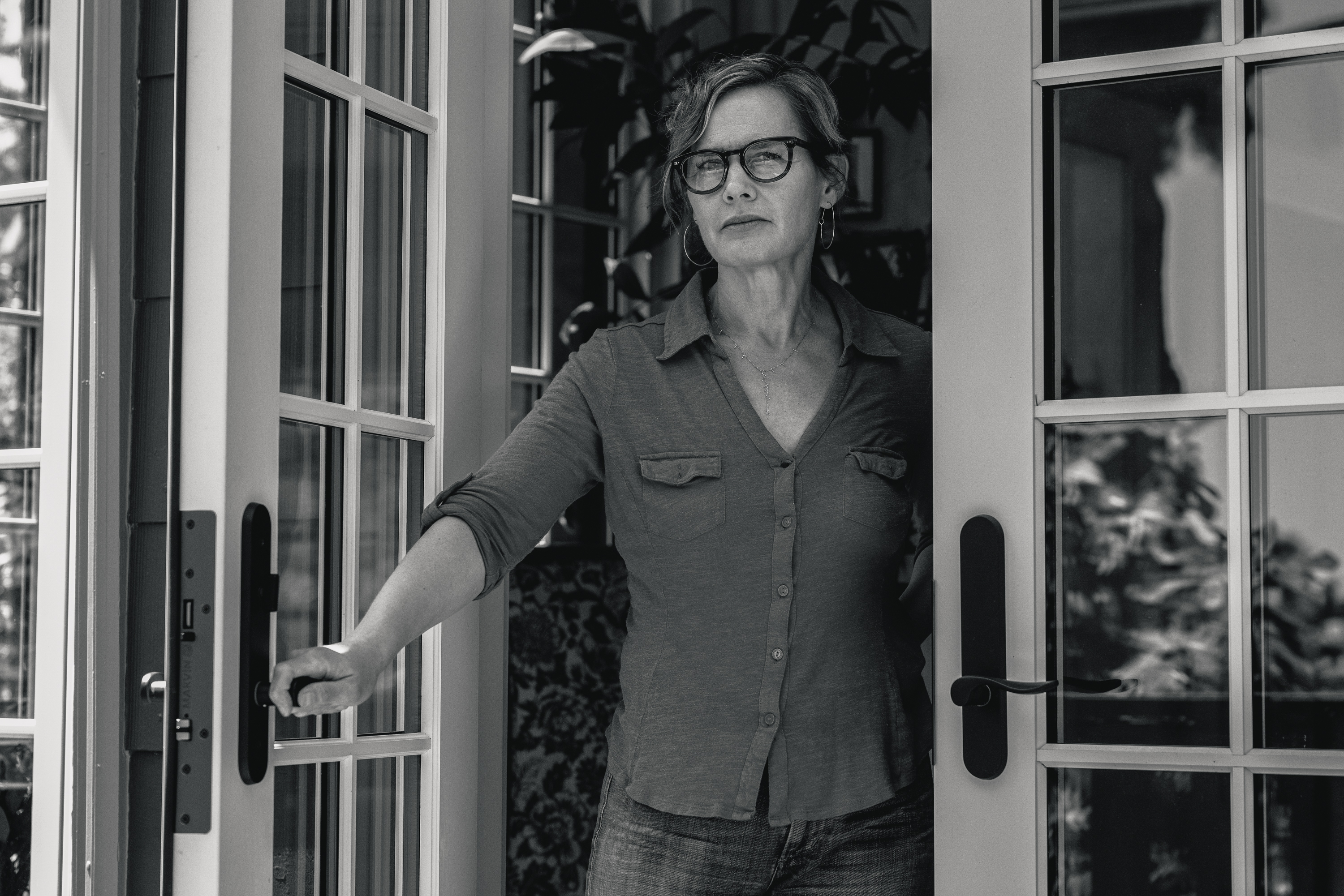 A woman with short hair and glasses stands in a doorway with glass panels. She is wearing a button-up shirt, and she holds onto one of the door handles while looking outside.