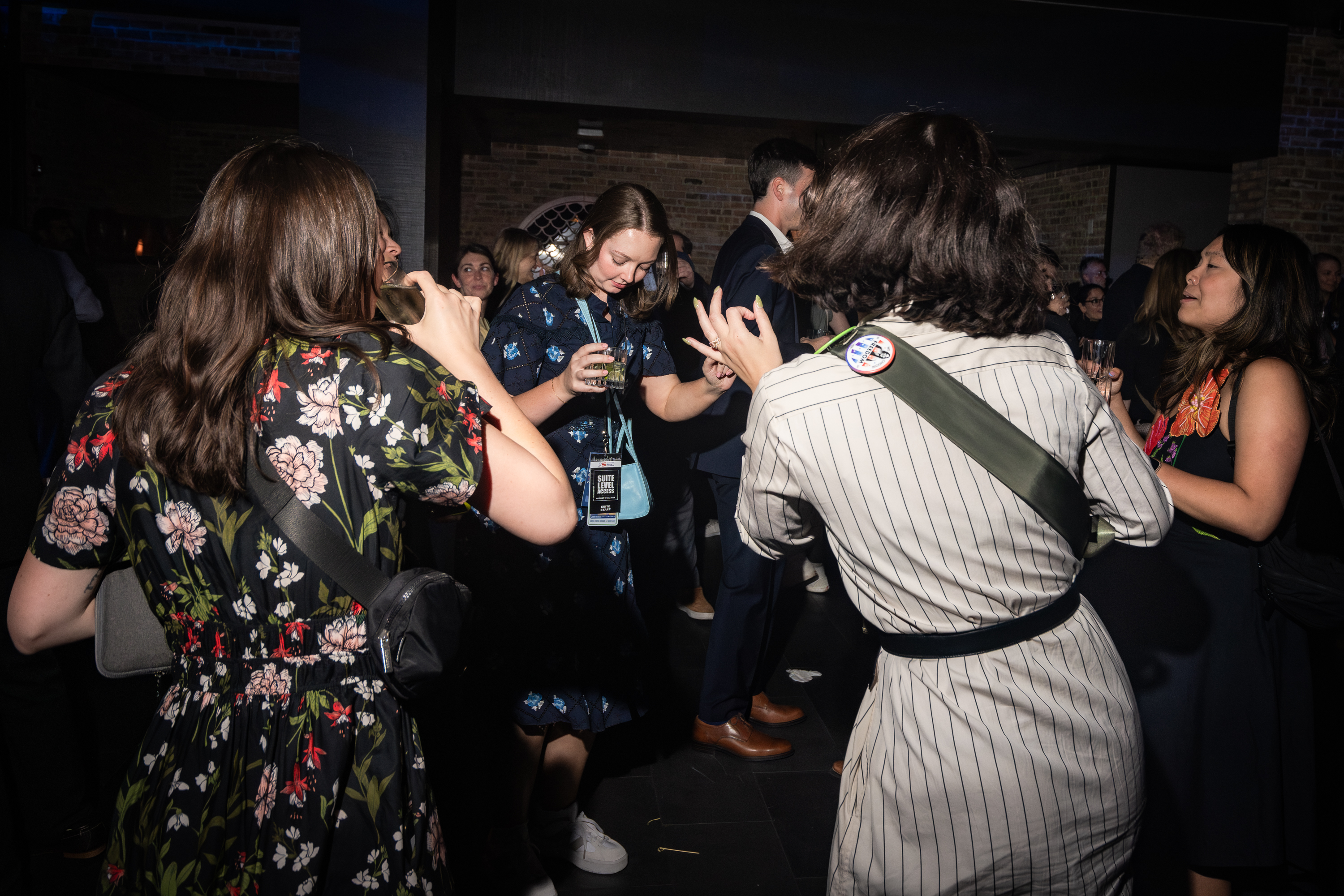 People in patterned dresses are dancing and socializing at a dark, lively event. Most hold drinks while chatting, and the background shows a crowded, brick-walled venue.