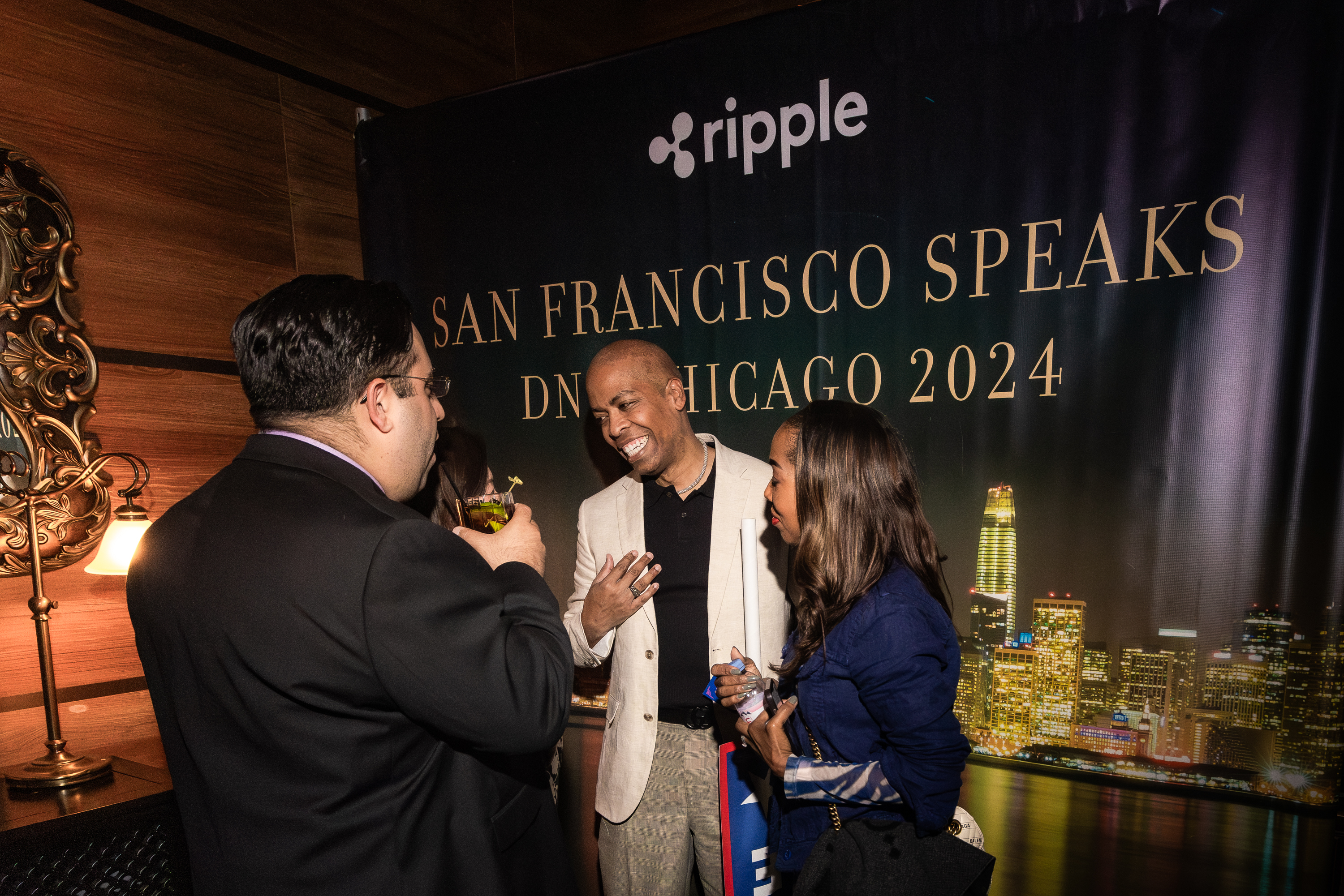 Three people are conversing cheerfully at an event with a backdrop that reads &quot;San Francisco Speaks DN Chicago 2024,&quot; with the logo for Ripple and a nighttime cityscape.