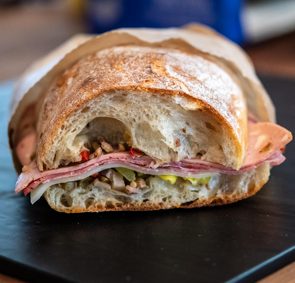 A sandwich with crusty bread, various types of sliced deli meat, and a filling of chopped vegetables and olives is shown, partially wrapped in paper.