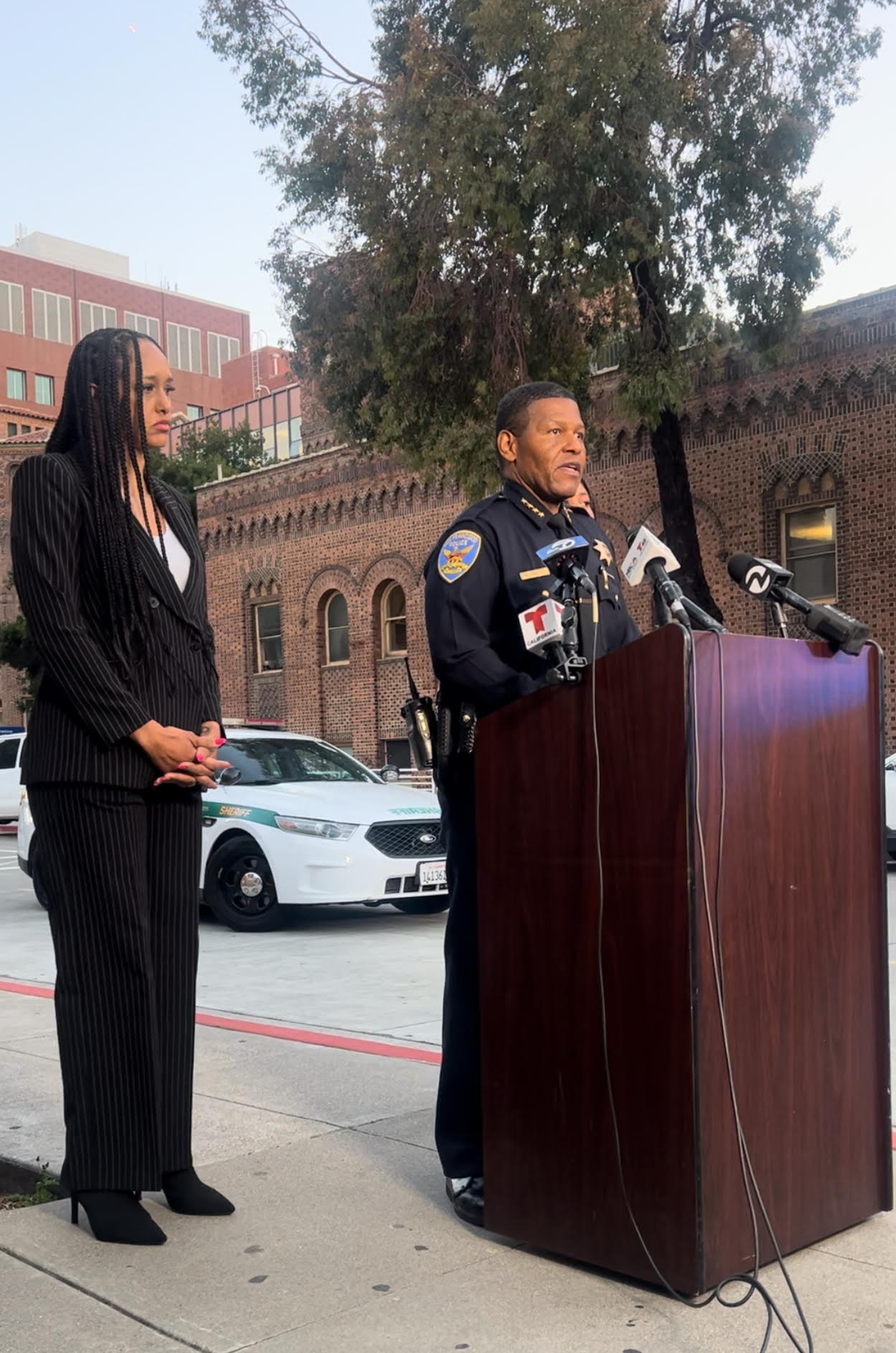 A police officer speaks at a podium with microphones outside a brick building, while a woman in a black pinstripe suit stands beside him. A police car is visible in the background.