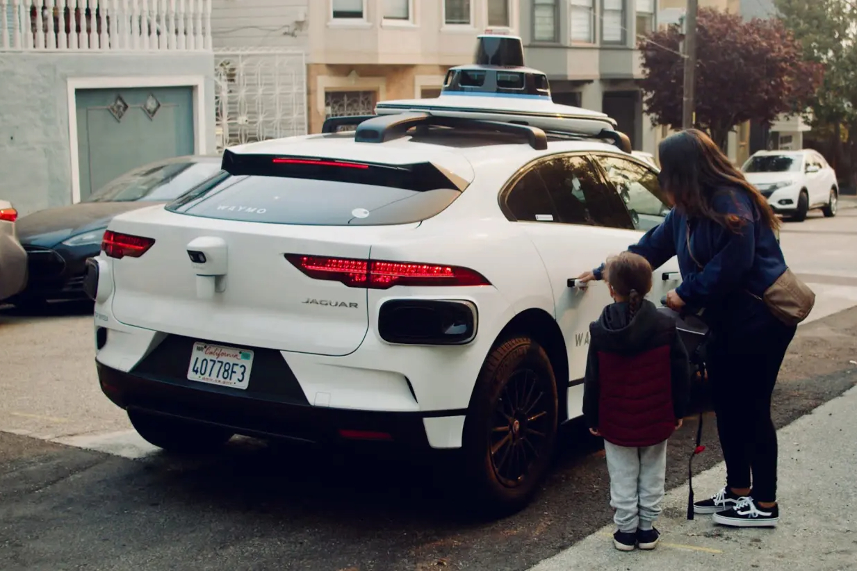 A woman and a child are seen on a street, with the woman opening the rear door of a white self-driving car equipped with sensors on its roof, labeled as a Waymo vehicle.