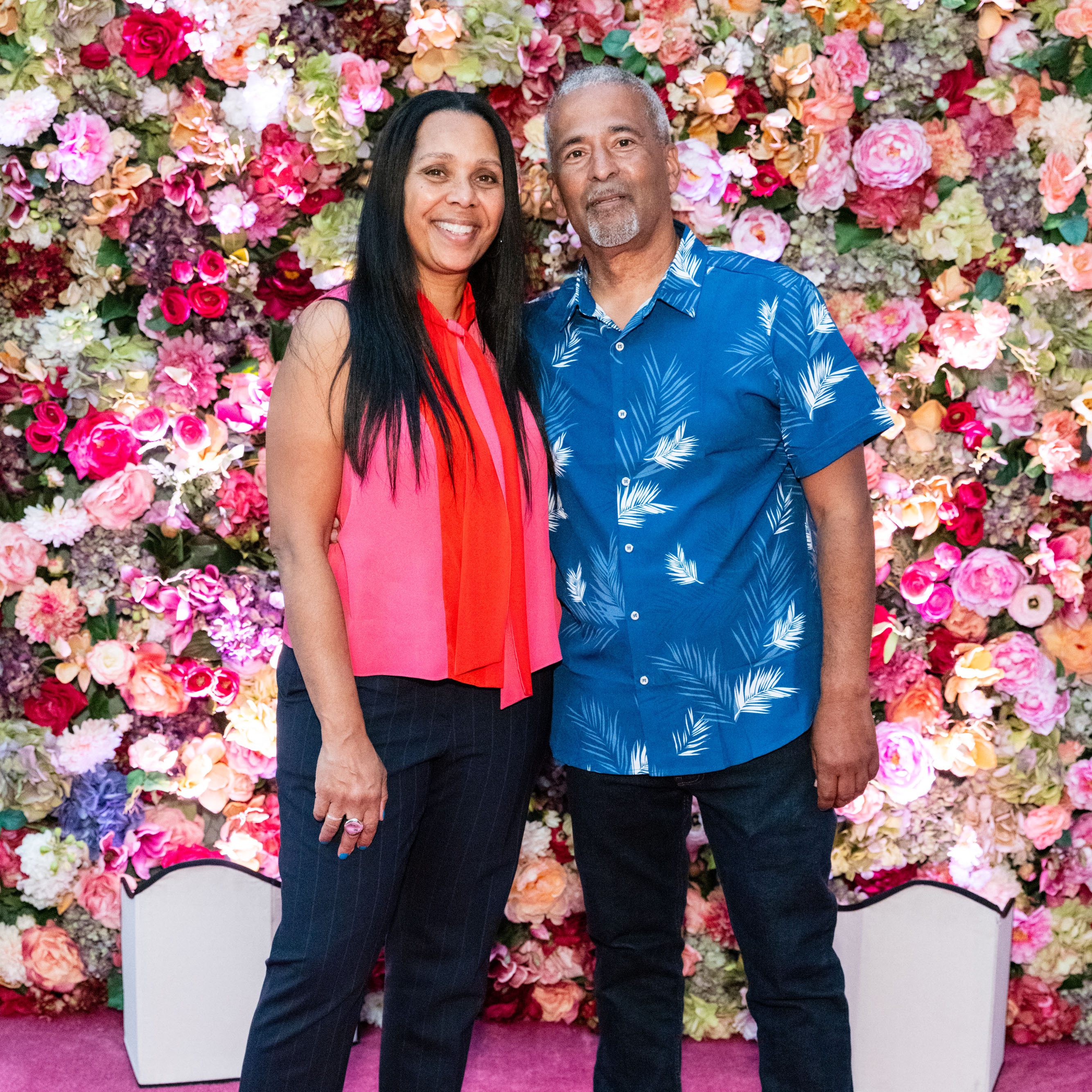 A smiling man and woman stand together against a vibrant, colorful flower wall. The woman wears a red sleeveless top, and the man wears a blue shirt with white leaf prints.