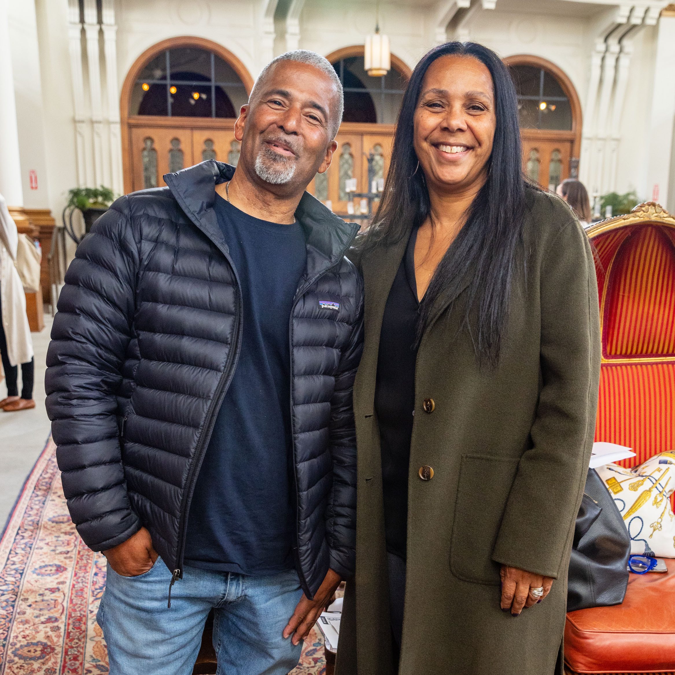 A smiling man in a black puffer jacket and jeans stands next to a smiling woman in a green coat in an ornately decorated room with arched doorways in the background.