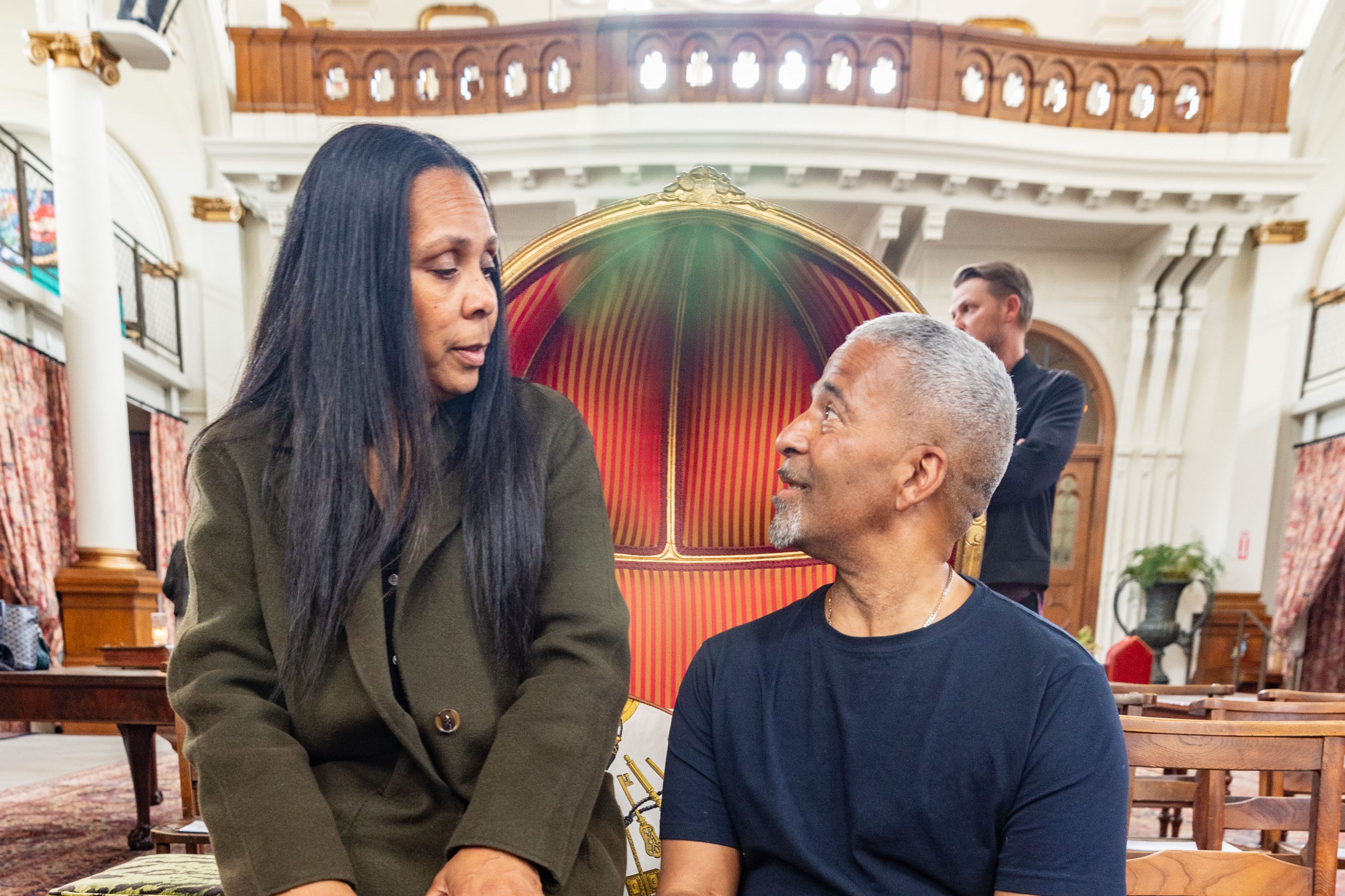 A woman and a man are talking and sitting in an ornate room with colorful rugs. The man is on a carved chair, while the woman, in a dark coat, perches on its arm.