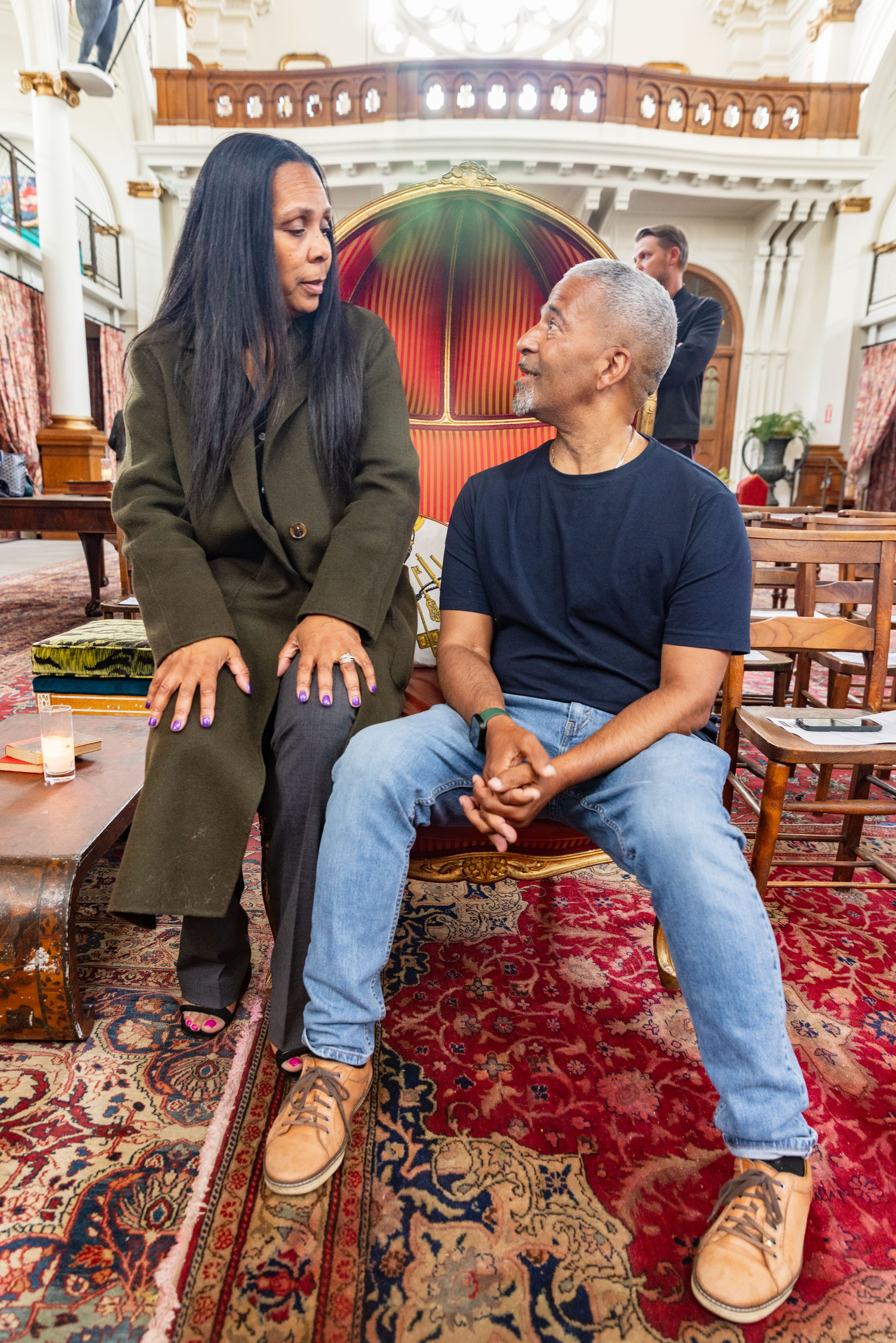 A woman and a man are talking and sitting in an ornate room with colorful rugs. The man is on a carved chair, while the woman, in a dark coat, perches on its arm.