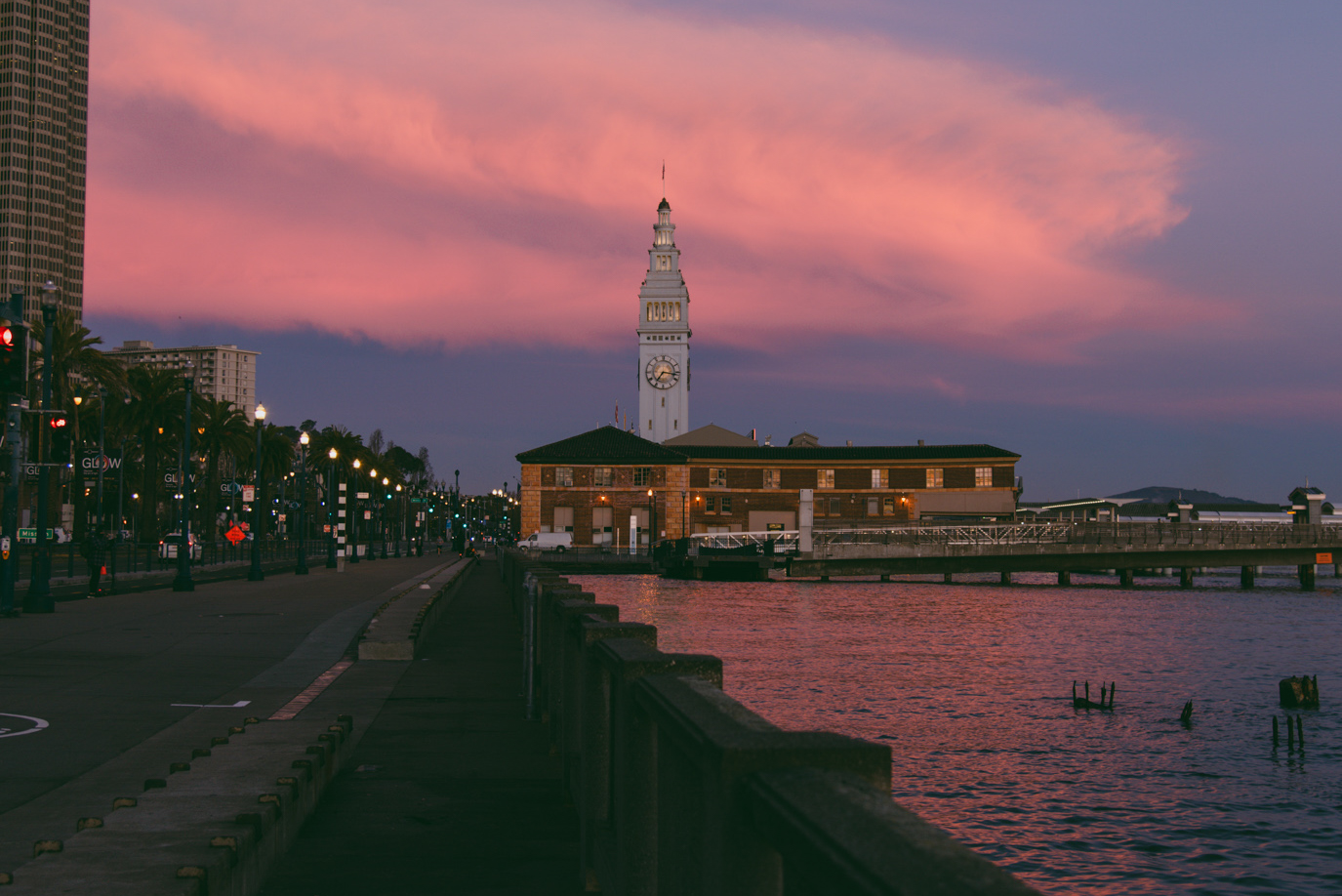 A clock tower overlooks a calm waterfront at dusk with a pier extending into the water and a row of streetlights illuminating a pathway lined with palm trees.