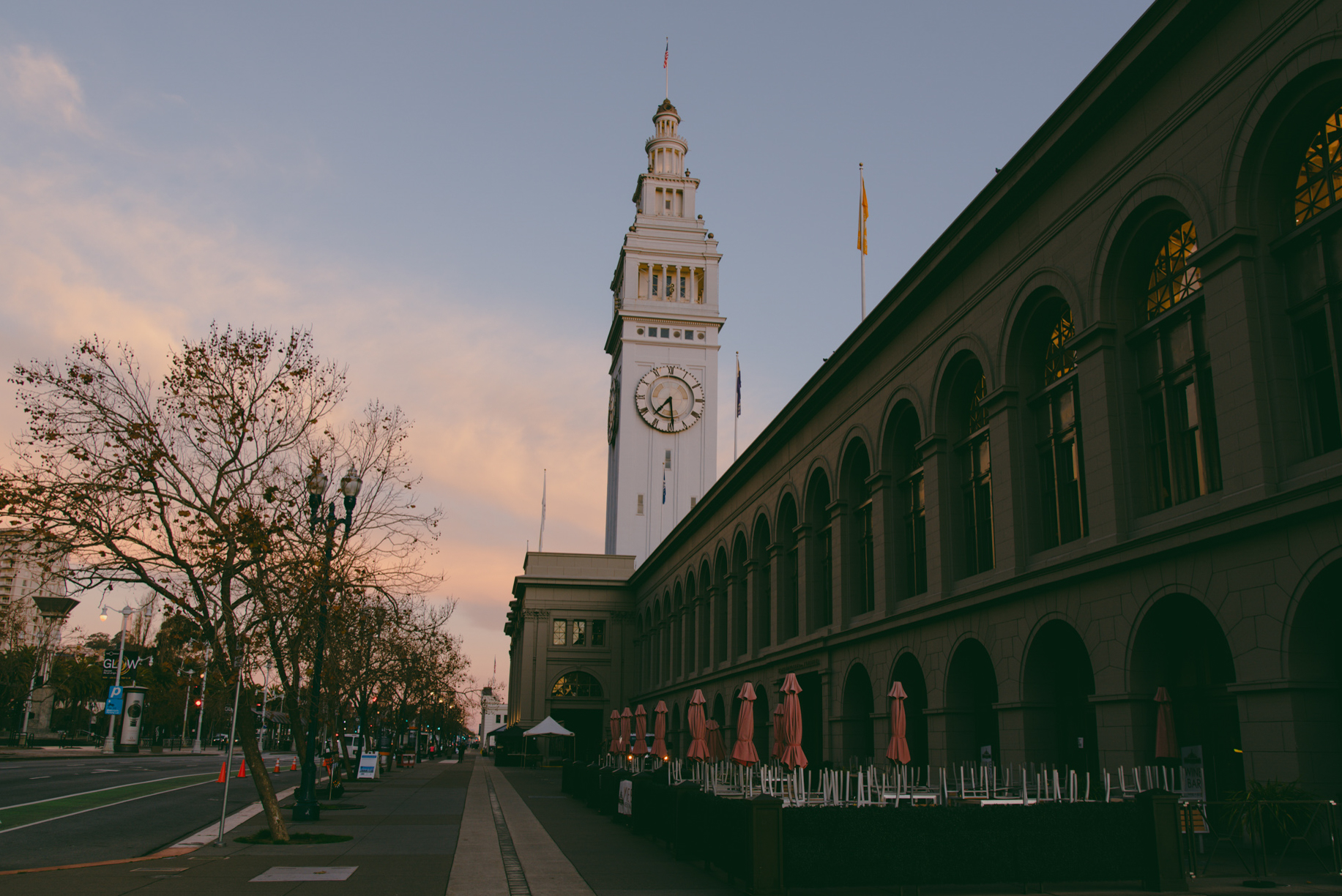 A tall clock tower stands against a pinkish sky. Adjacent is a long, arched building with stacked chairs and closed umbrellas outside on a quiet street.