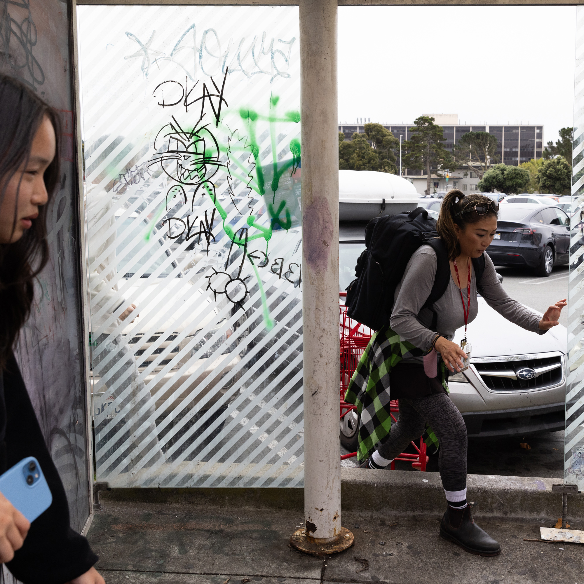 A woman with a backpack steps over a ledge next to a graffitied panel, while another person nearby holds a blue phone. Cars and a building are in the background.