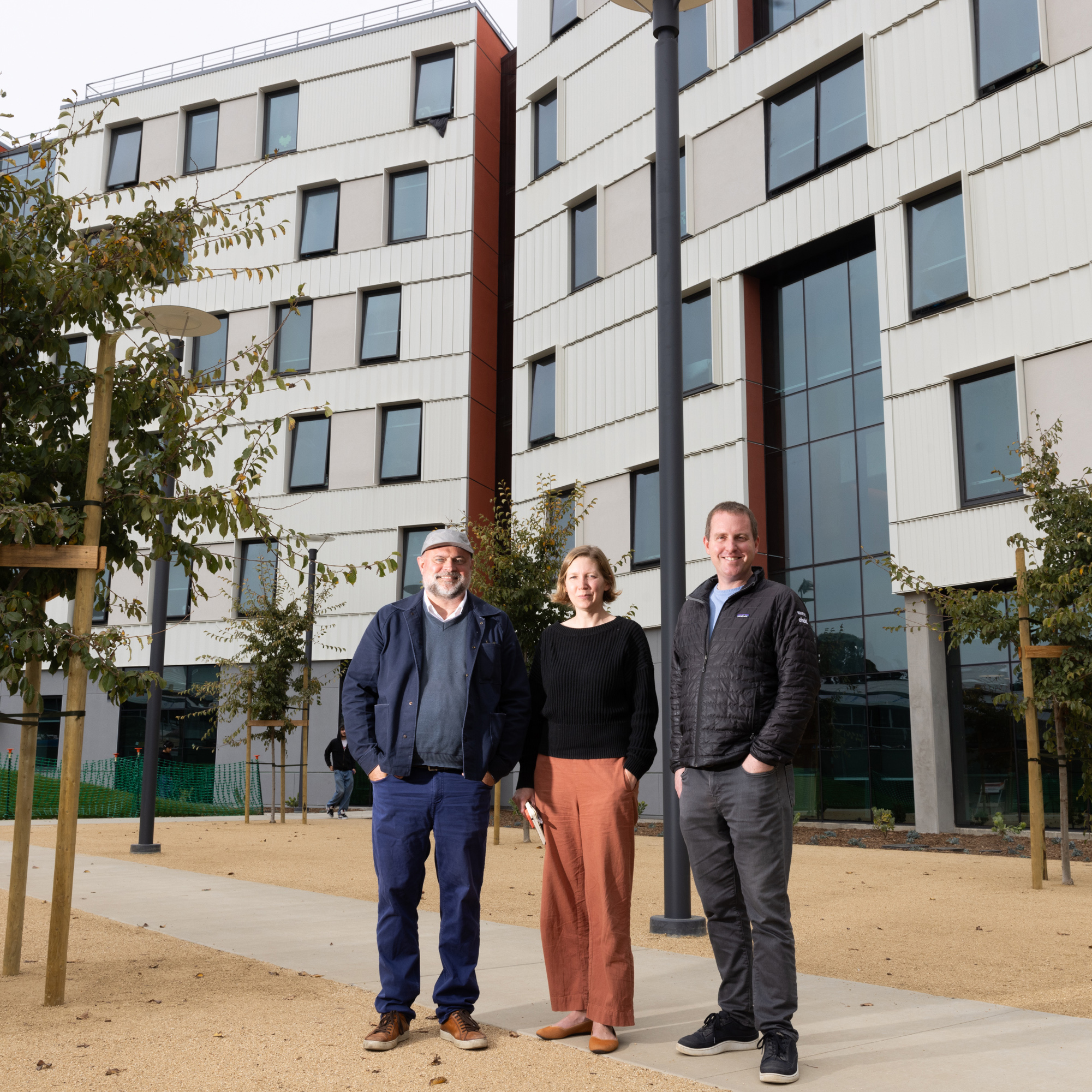 Three people stand on a pathway outside a modern, angular building with large windows and trees lining the walkway.