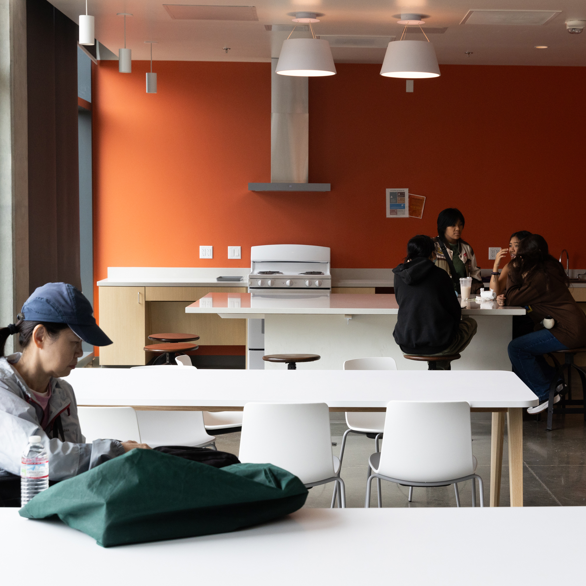 A bright room with orange walls, white tables, and modern pendant lights features one person alone in the foreground and four people conversing in the background kitchen area.