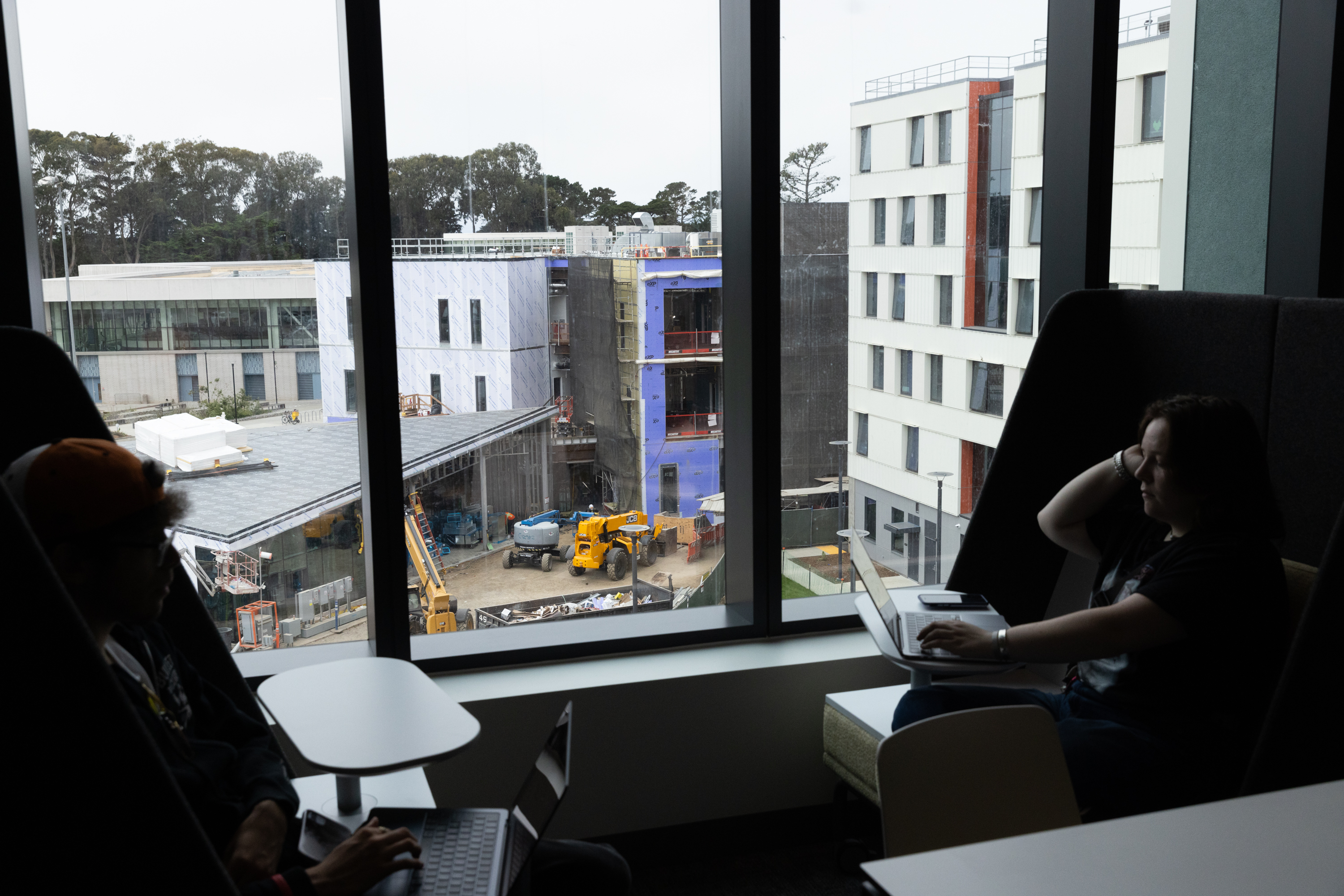 Two people work on laptops by large windows overlooking a construction site with machinery and multiple buildings, surrounded by trees in the background.