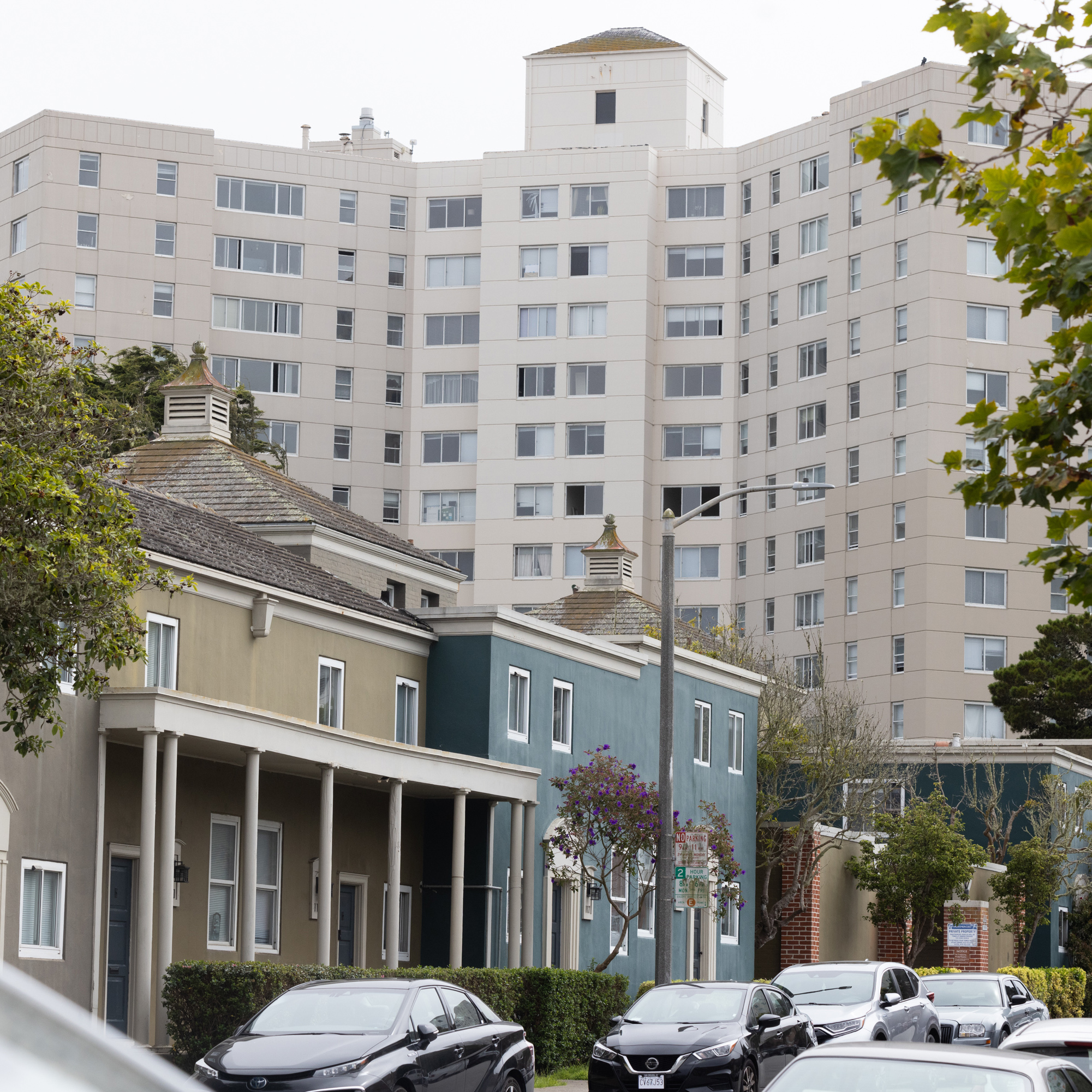 The image shows modern townhouses with green and teal facades in front of a large beige apartment building. Cars are parked along the street.