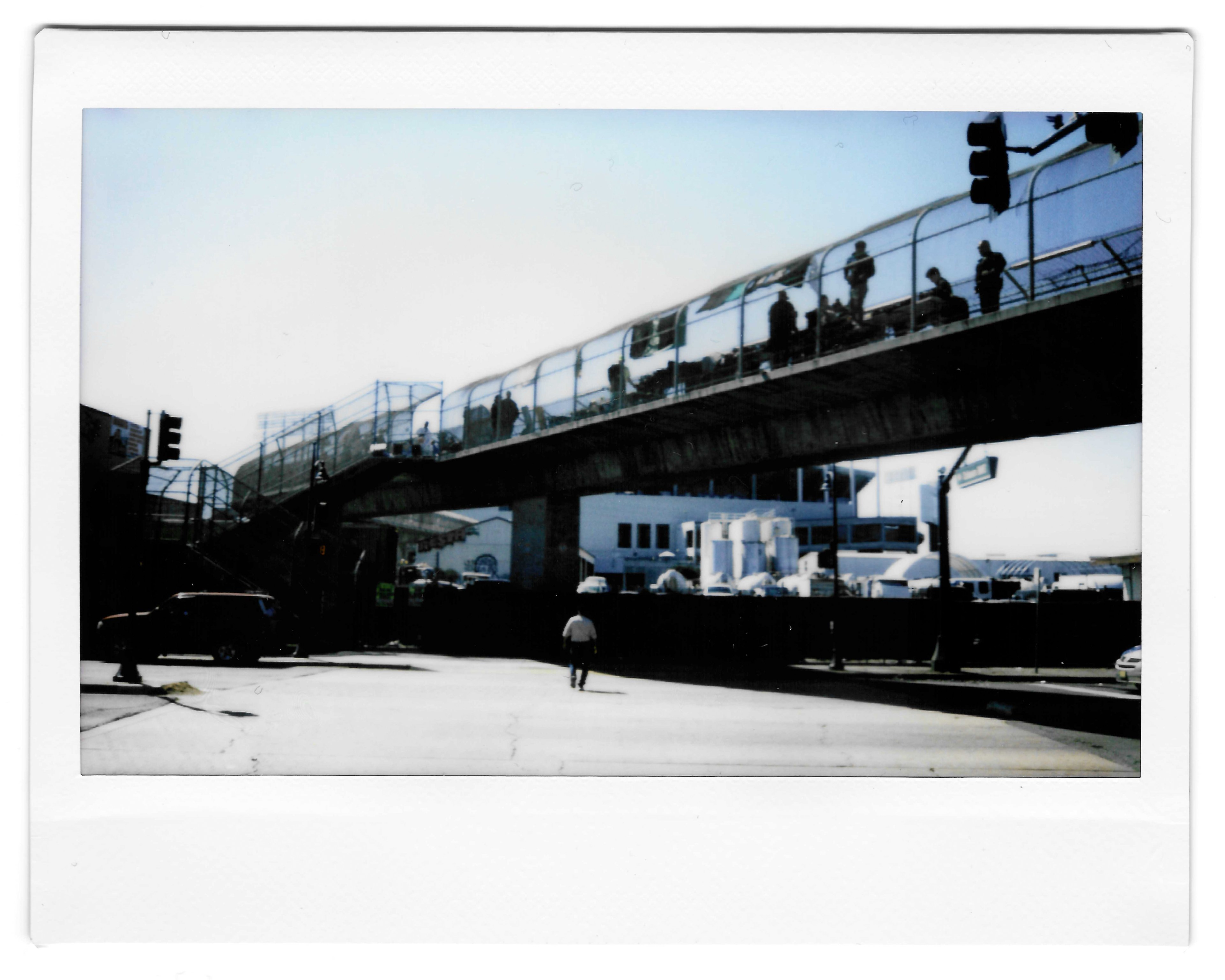 The image shows a pedestrian bridge over a street, with people walking on it. Below, a lone person walks toward the bridge, and there are buildings and cars nearby.