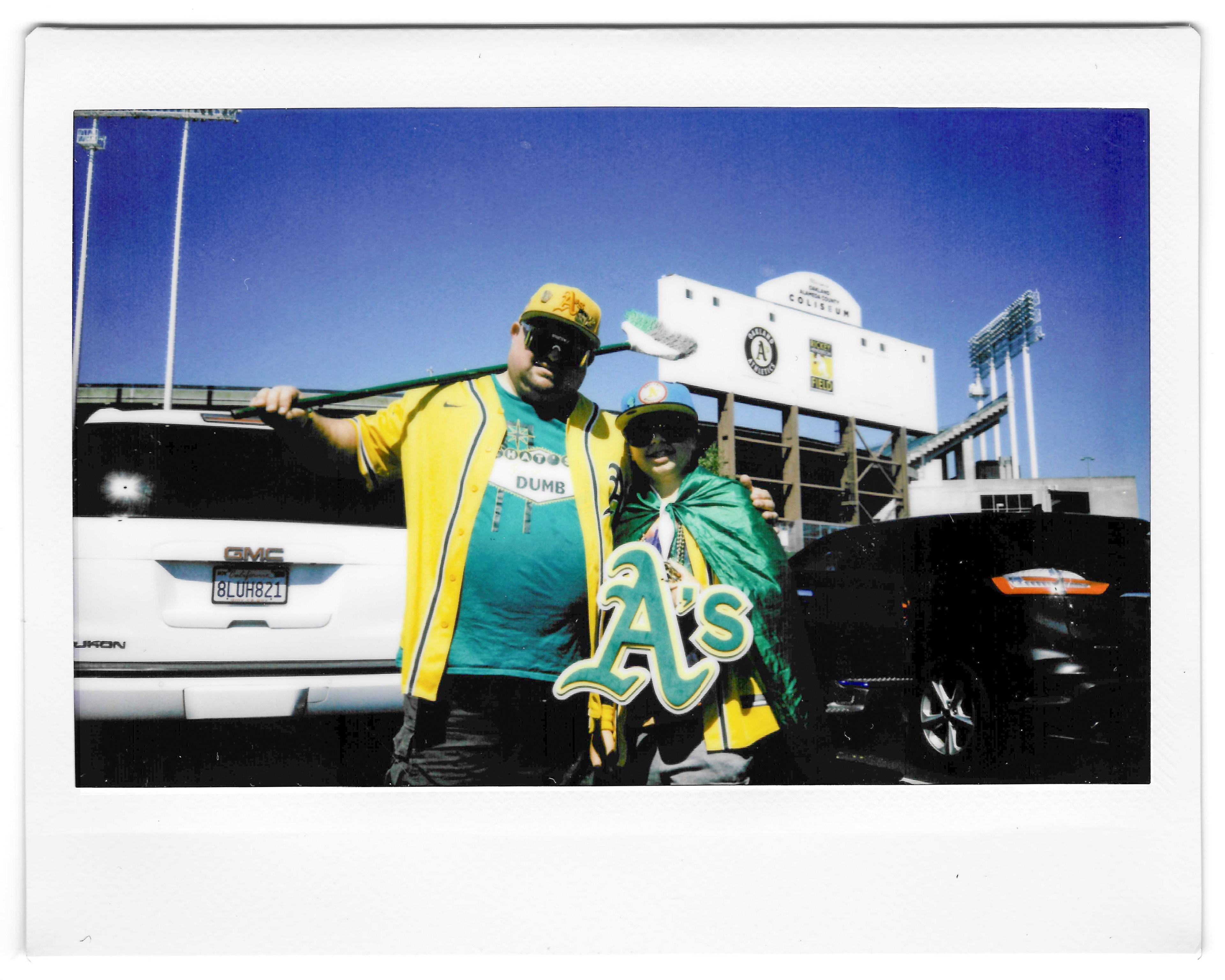 Two people, dressed in Oakland Athletics gear, pose in a stadium parking lot. One holds an &quot;A's&quot; sign, with the Oakland Coliseum visible in the background.