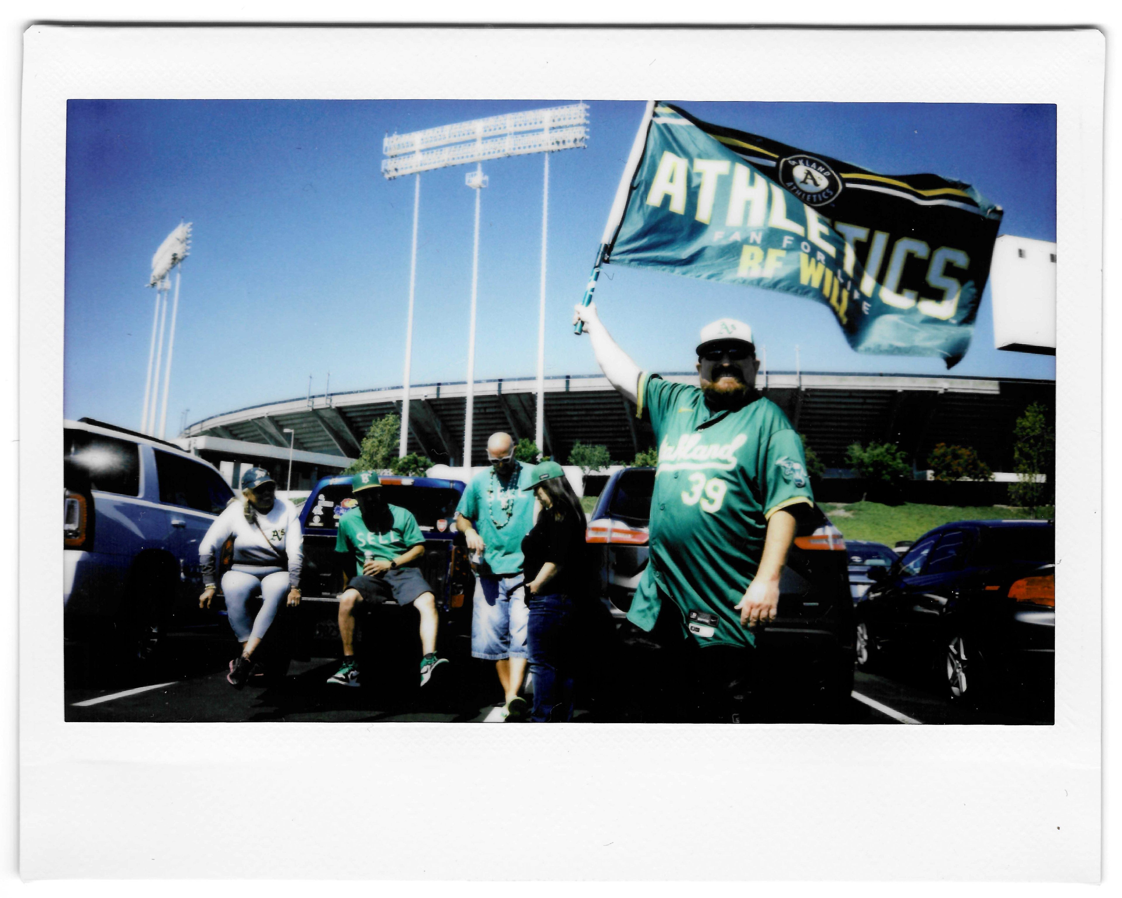 A group of fans in Oakland Athletics attire stands and sits by cars in a parking lot, with one man holding a large Athletics flag. The stadium is visible in the background.