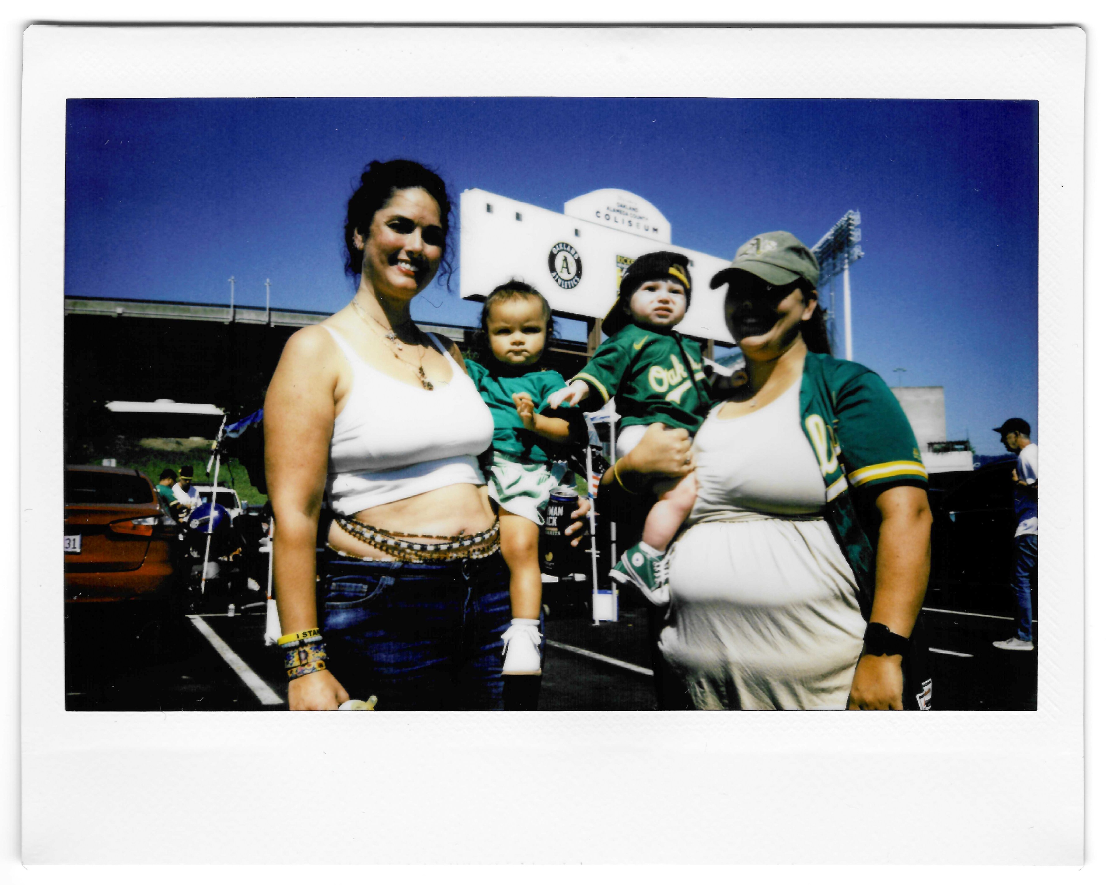 Two women are posing with two babies in a parking lot. One woman wears a white tank top and blue jeans; the other wears an Oakland A's jersey and cap.