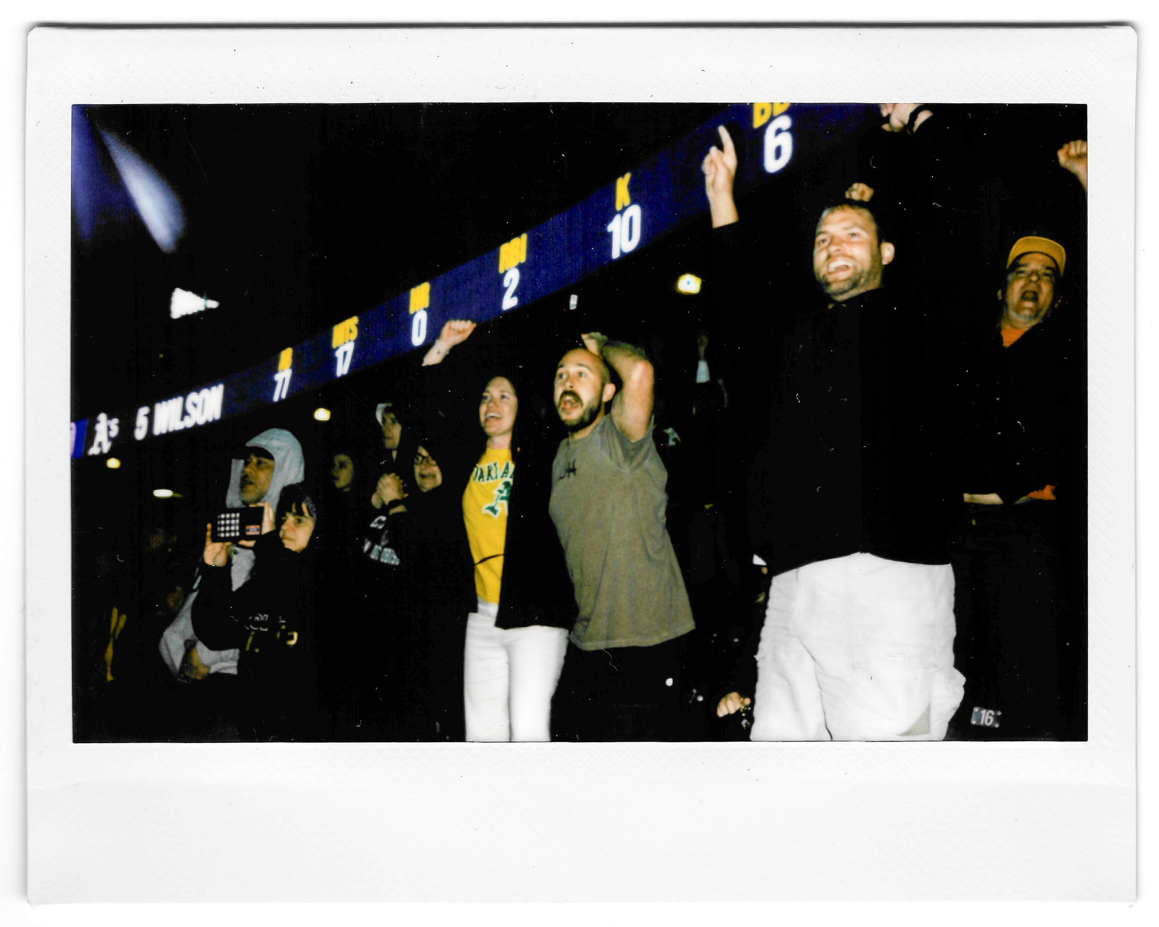 A group of excited fans, some with raised arms, are captured in front of a scoreboard displaying names and numbers. The scene suggests a lively sports event.