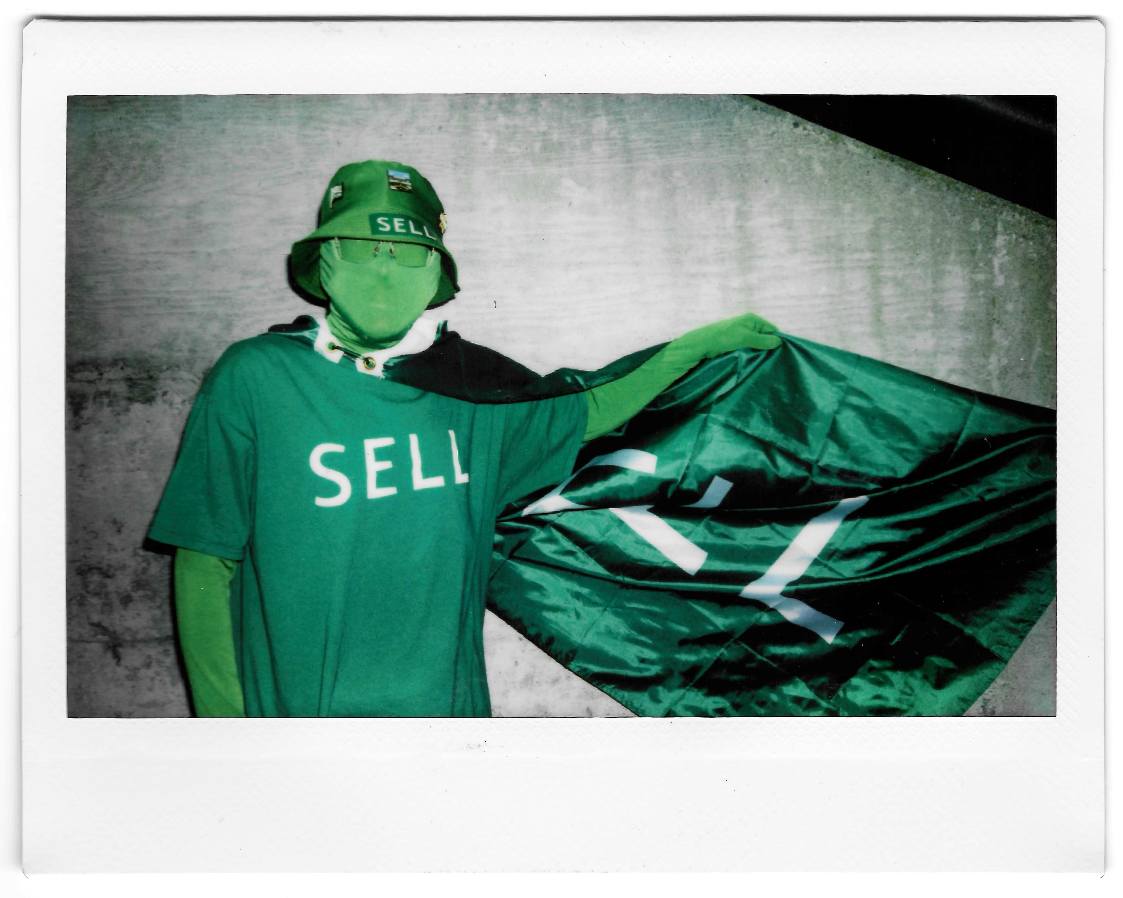 A person wearing a green bodysuit, &quot;SELL&quot; t-shirt, and bucket hat holds a green flag with white symbols. The background is a plain, grey wall.