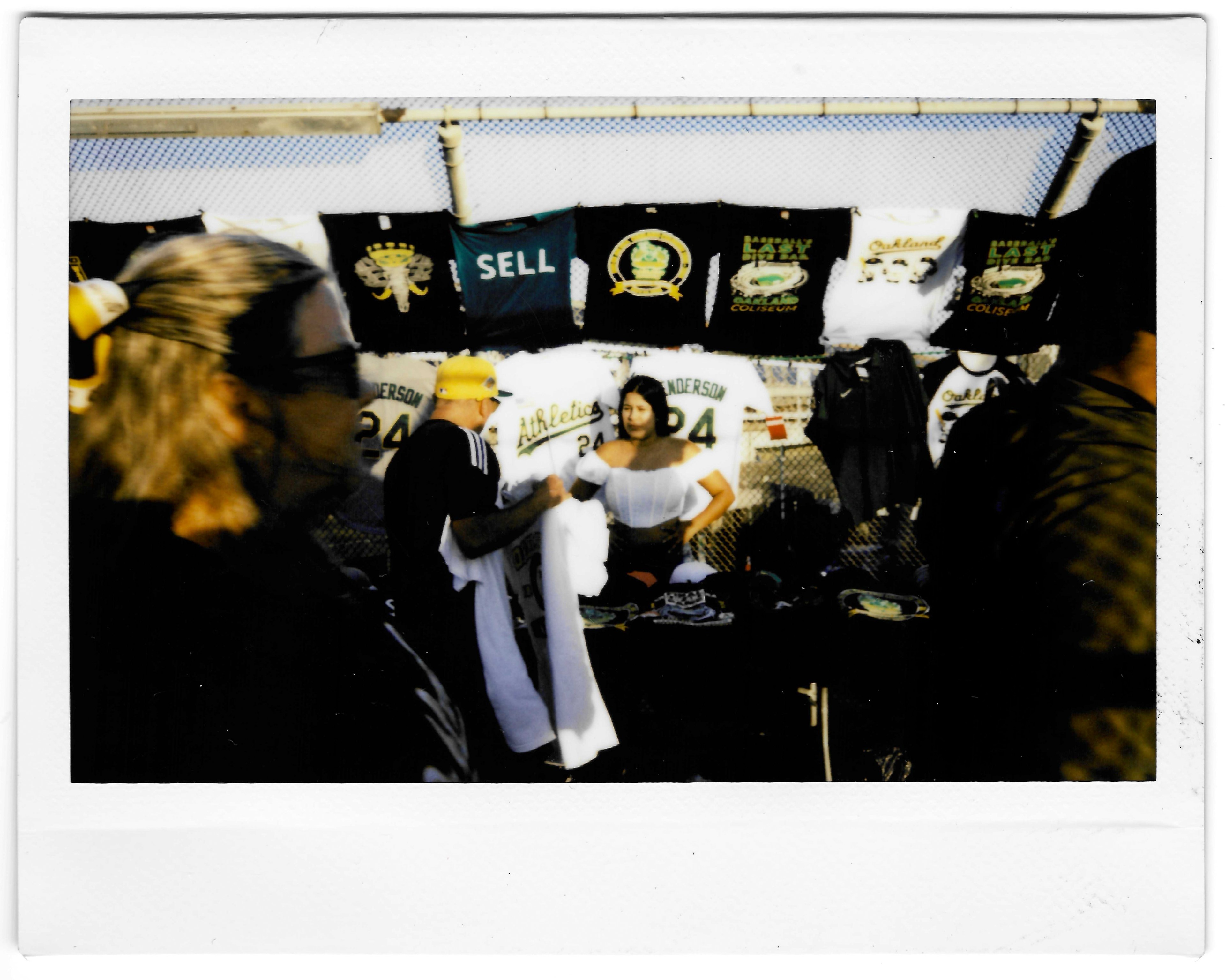 People are gathered at an outdoor merchandise stall, with various T-shirts hanging on a fence in the background. Two people are in the foreground, with one examining an item.