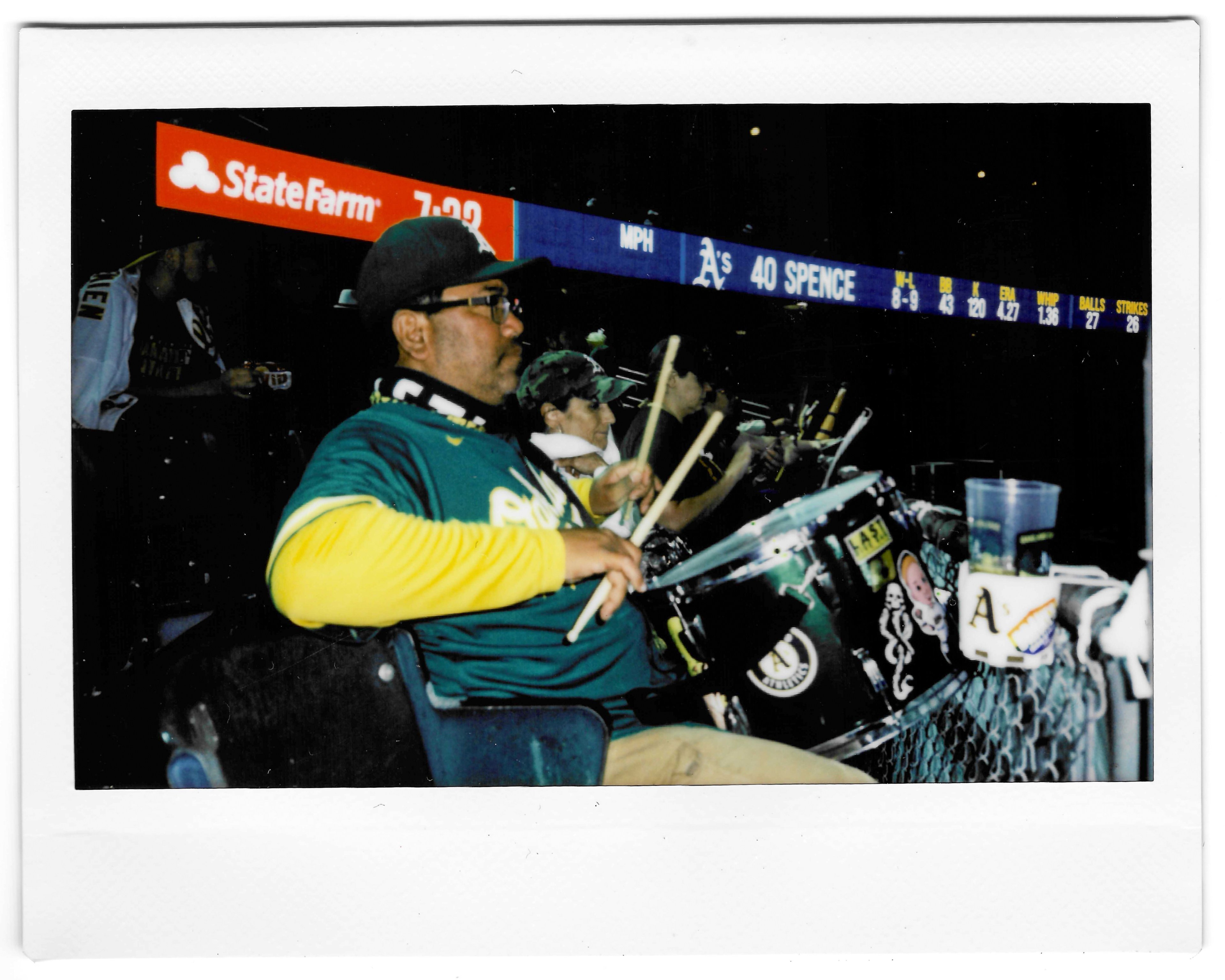 A group of enthusiastic fans, wearing green and yellow apparel, play drums in a sports stadium. A scoreboard behind them shows game statistics and an advertisement.