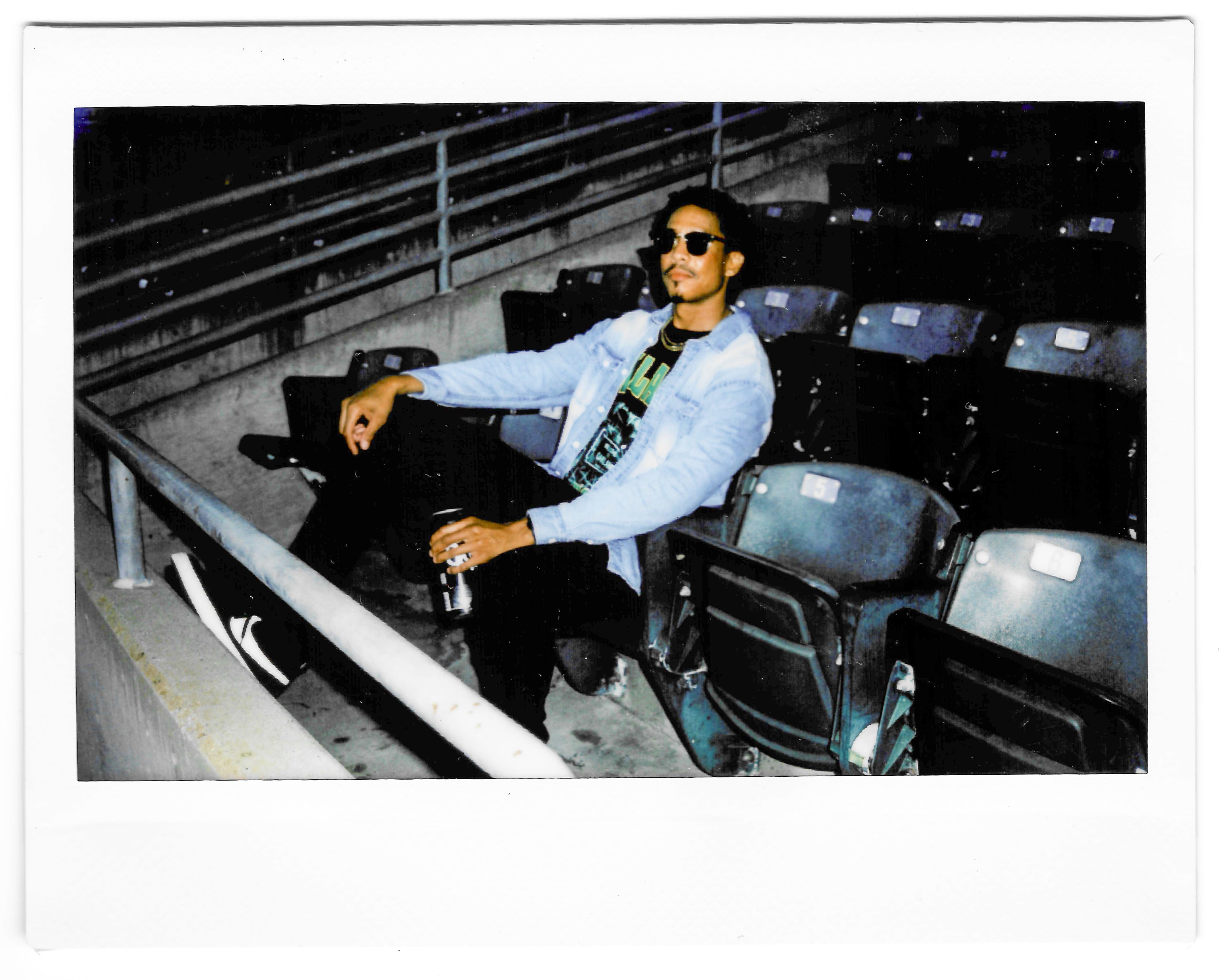 A person sits relaxed in an empty stadium seat, wearing sunglasses, a denim jacket, and black pants, holding a drink can, with one leg propped up on a railing.