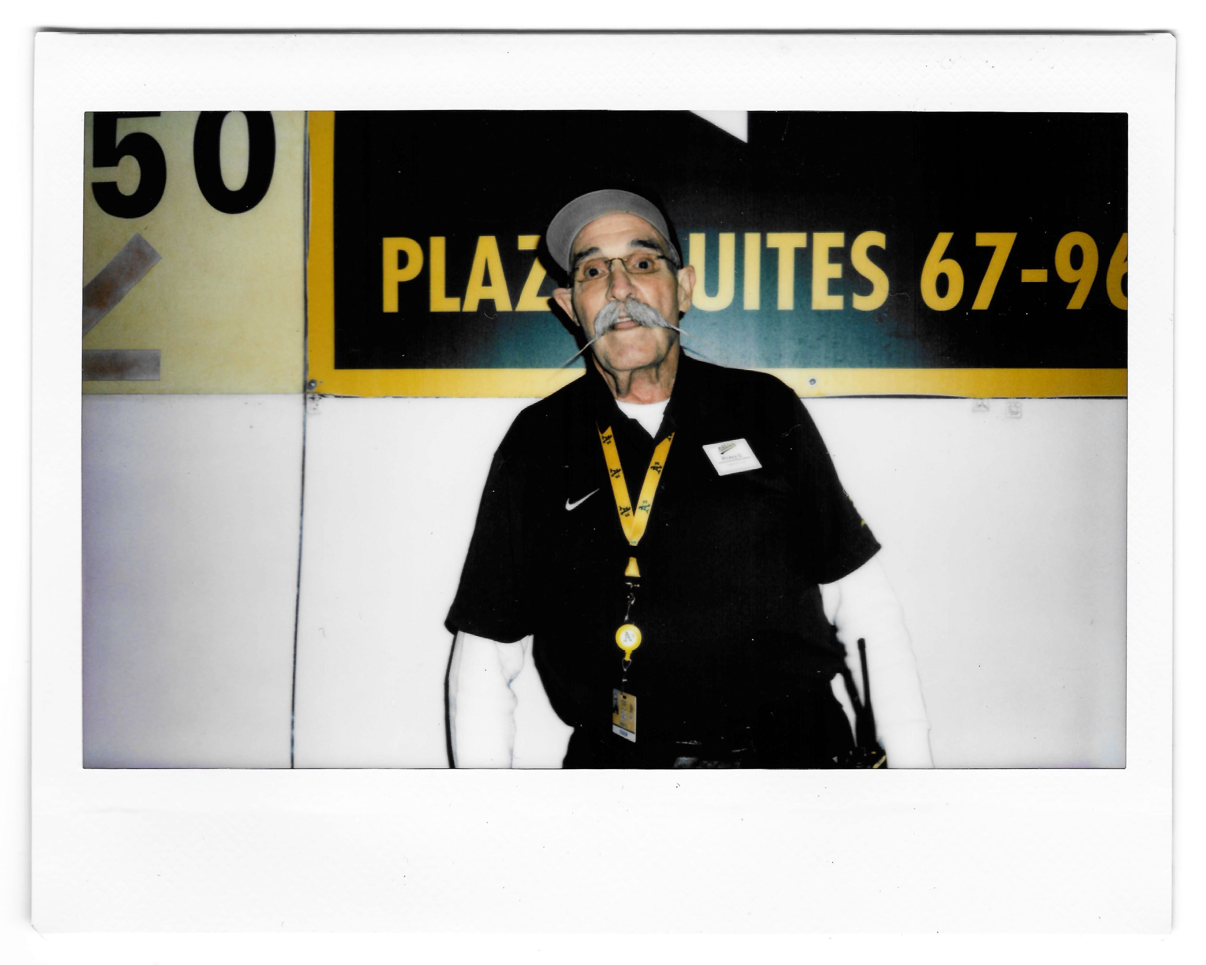 An older man with a gray cap, glasses, and a prominent mustache stands in front of a yellow and black sign. He wears a black shirt with a yellow lanyard and name badge.