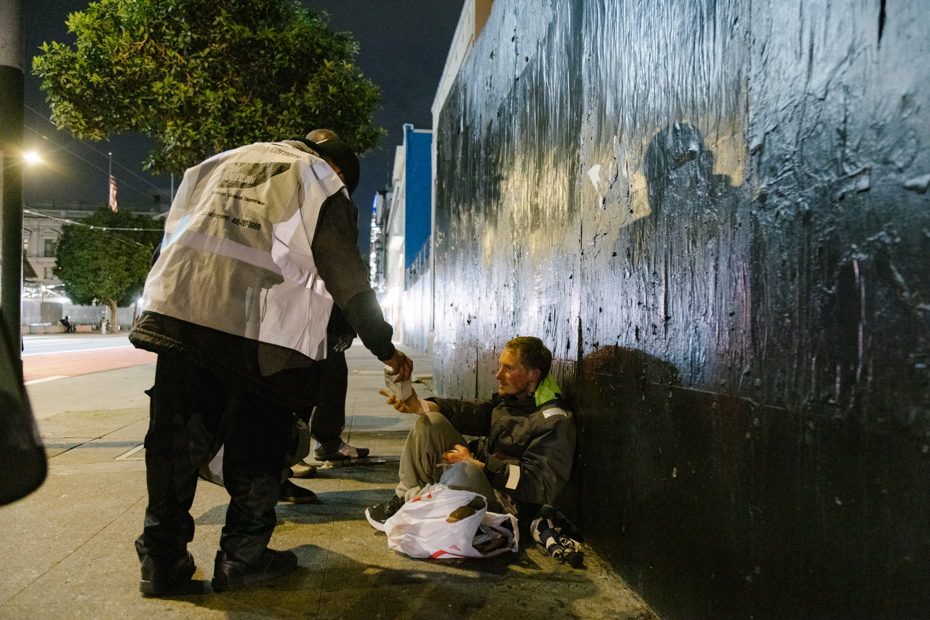 At night, a person in a white vest helps a seated man against a dark wall, handing him something. The seated man has bags near him, and a leafy tree is in the background.