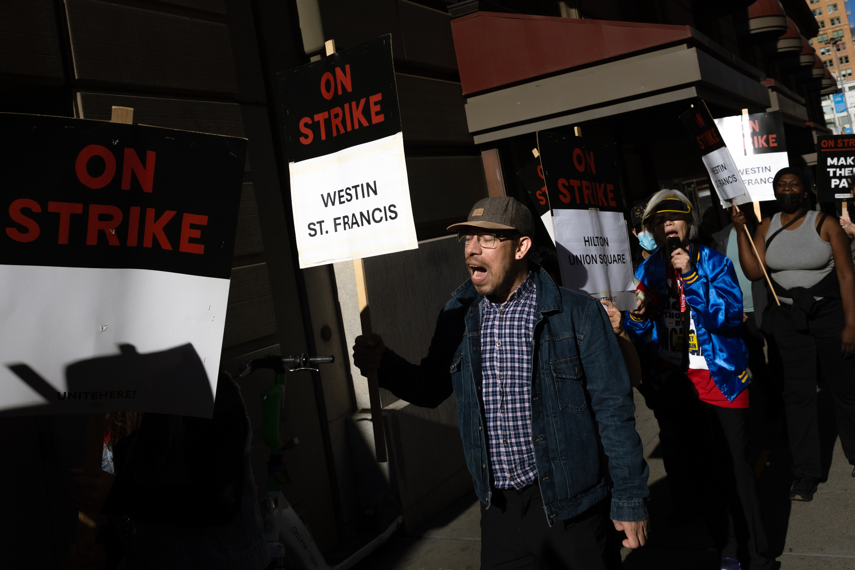 A group of people holding "On Strike" signs, including names like "Westin St. Francis" and "Hilton Union Square," are protesting on a city street.