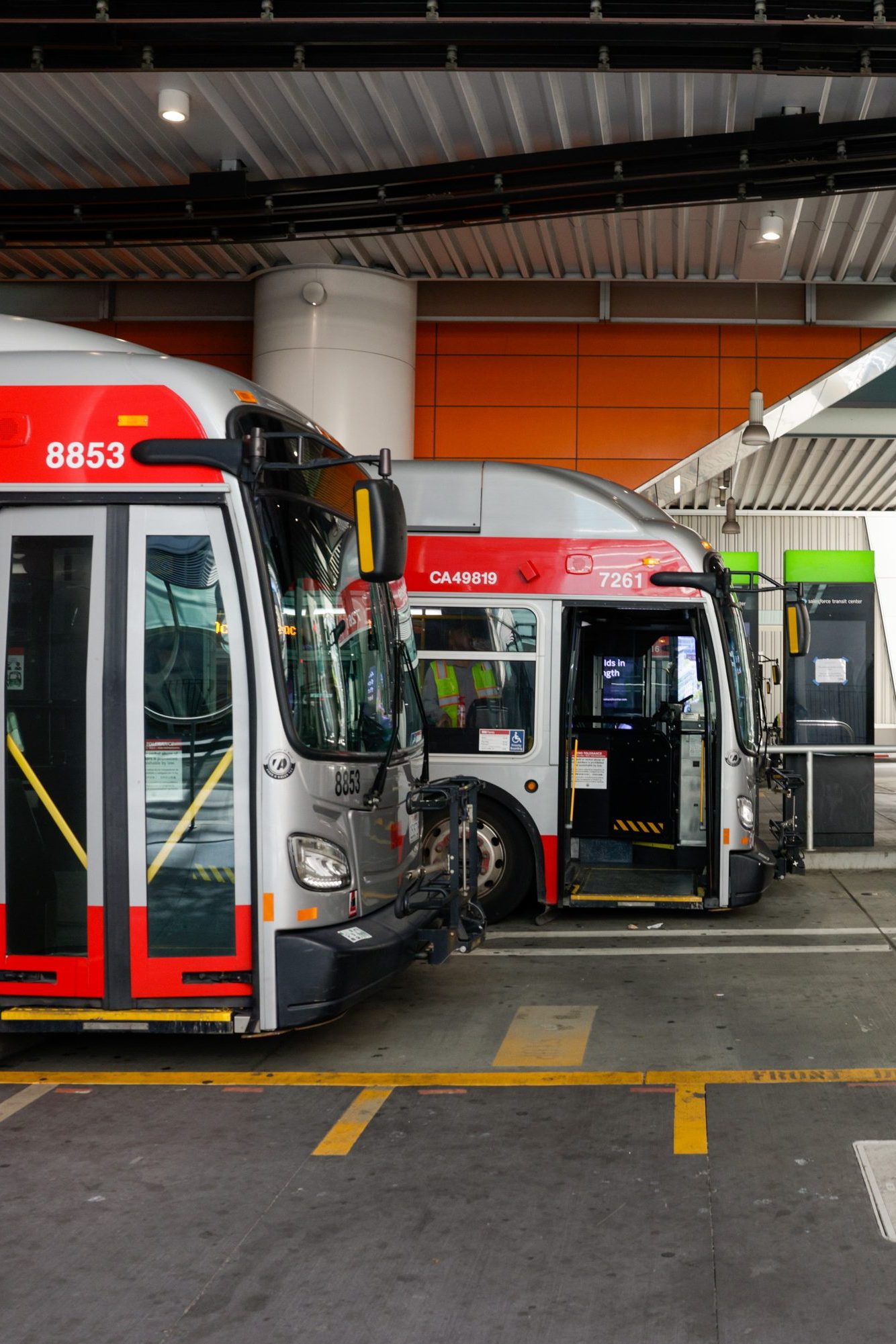 The image shows city buses parked in a well-lit, modern bus terminal with orange accents. Two people are seen walking nearby, and one bus has its doors open.